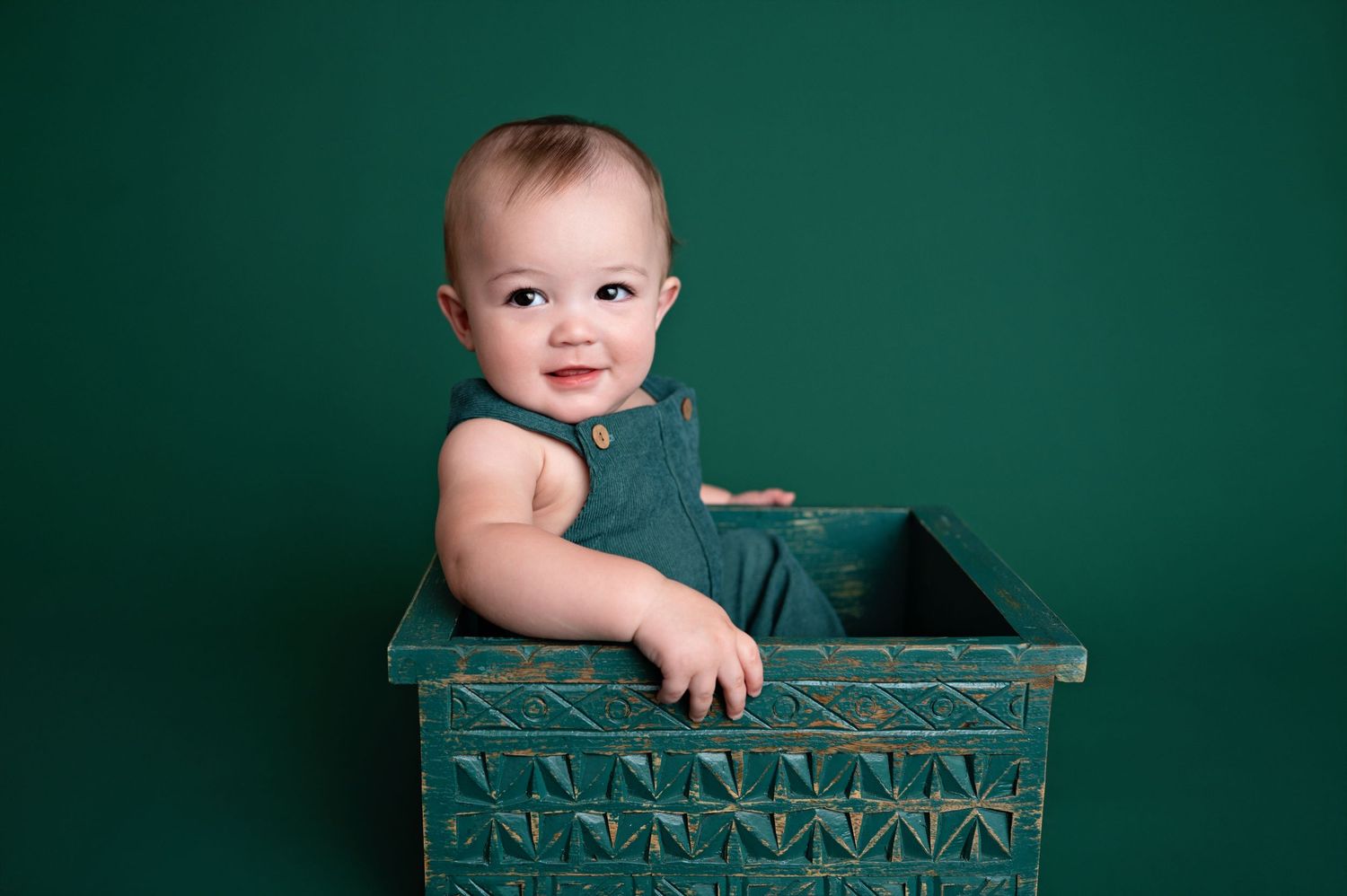 Small child wearing denim outfit sits in decorative turquoise wooden box against dark green background.