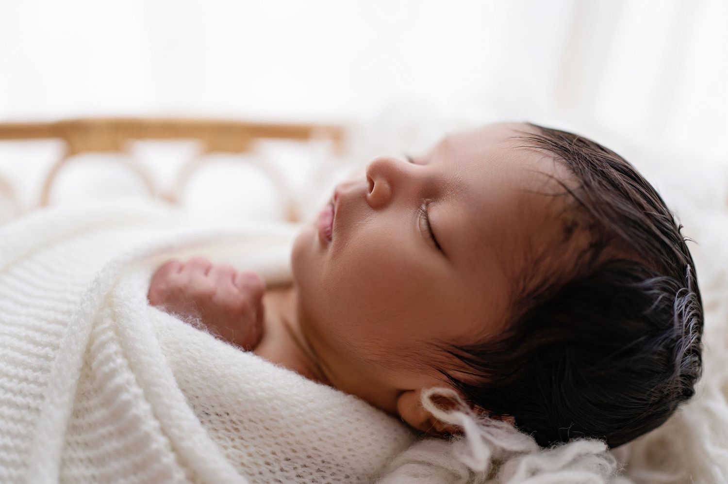 Peaceful newborn baby swaddled in white blanket resting on white textured surface.