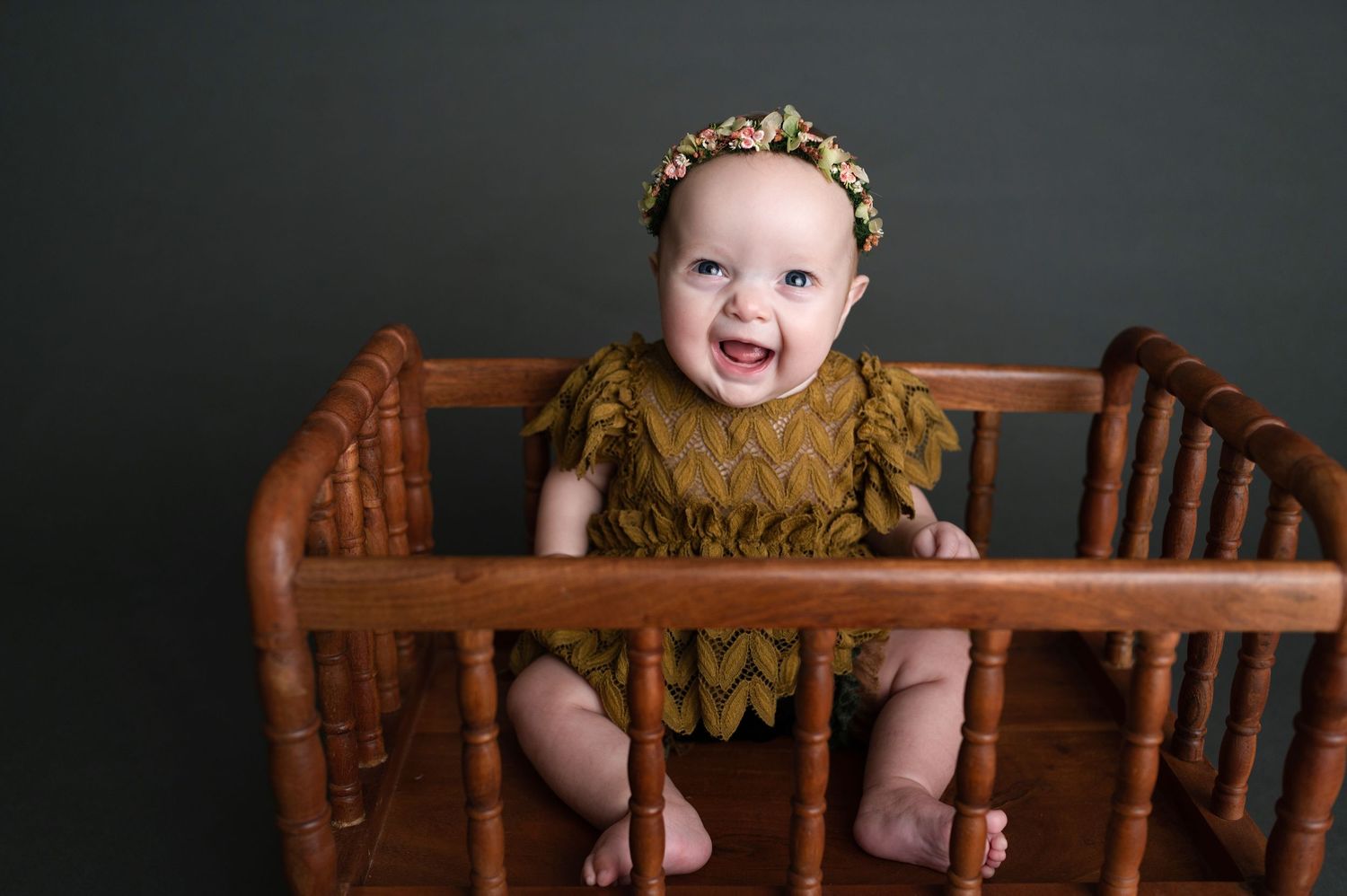 Baby in olive green outfit sits excitedly in wooden crib with gold floral headband.