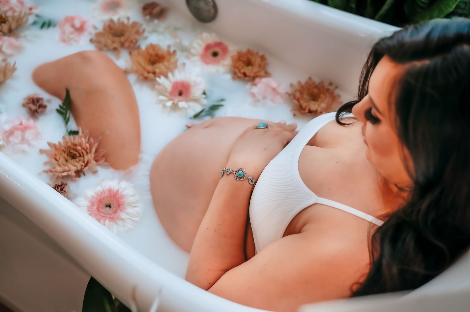 A pregnant woman relaxes in a bathtub filled with pink and peach flowers wearing a white top in a serene bath setting.