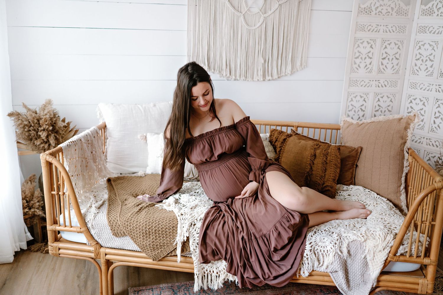 A brown off-shoulder maxi dress styled with boho decor and rattan furniture in a light-filled room.