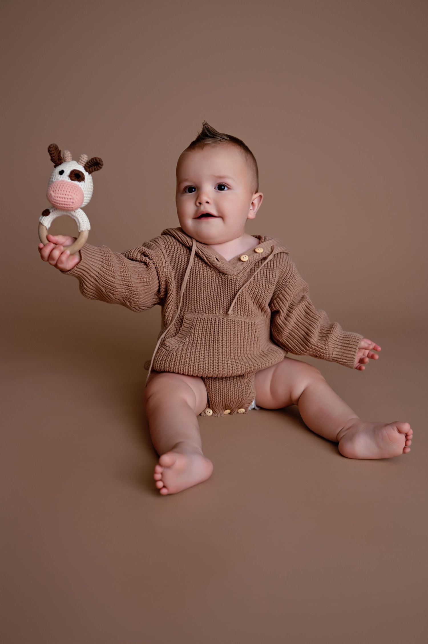 Baby wearing tan knit sweater poses playfully on brown backdrop in various positions with stuffed animal toy.