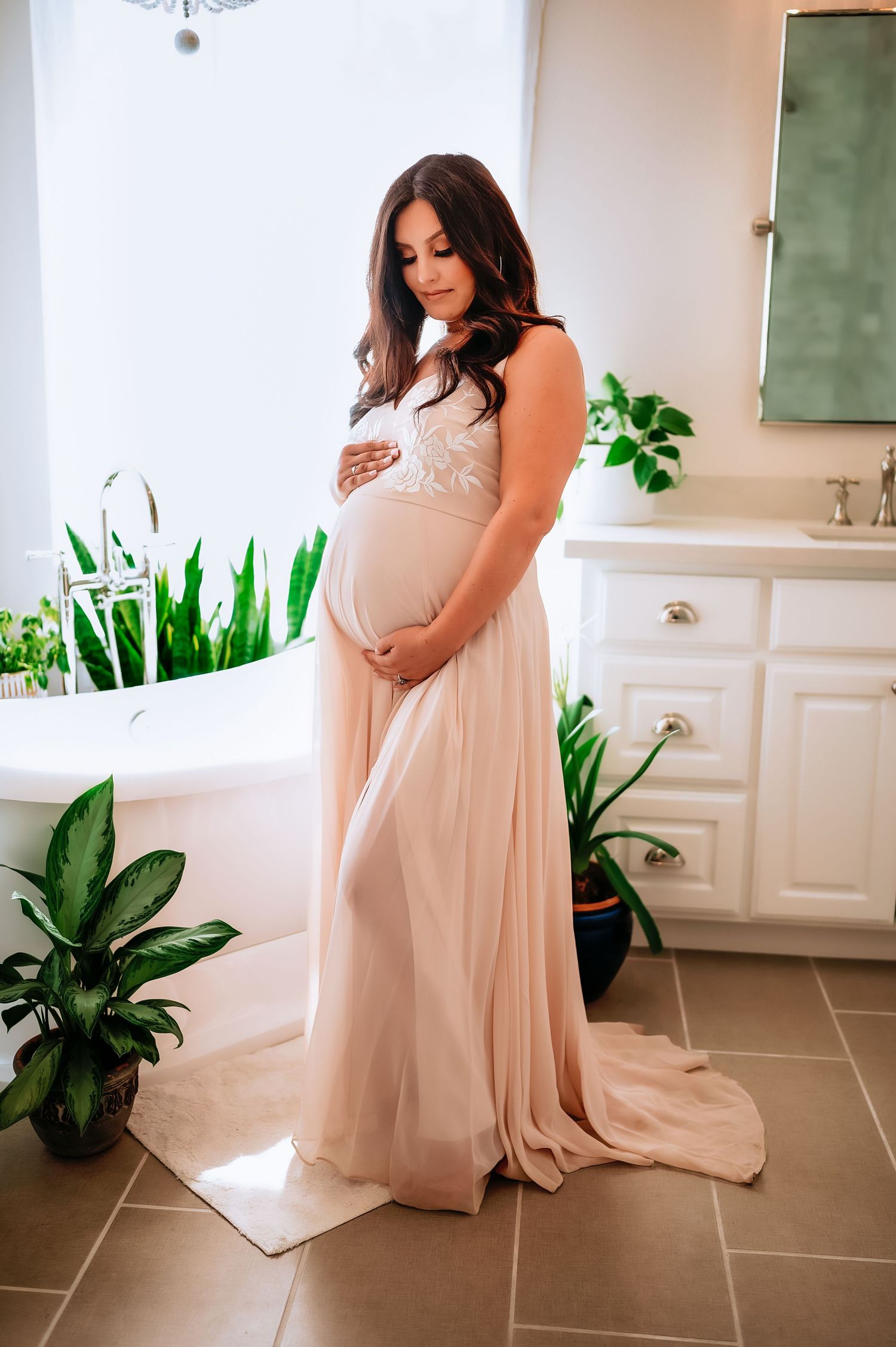 An expectant mother in a flowing blush gown stands in an elegant bathroom with indoor plants.