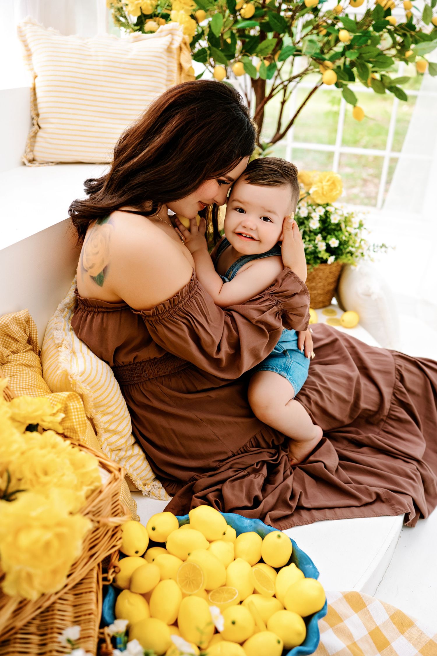 A mother in a brown dress cuddles her smiling baby boy surrounded by lemons and yellow flowers in a bright studio.