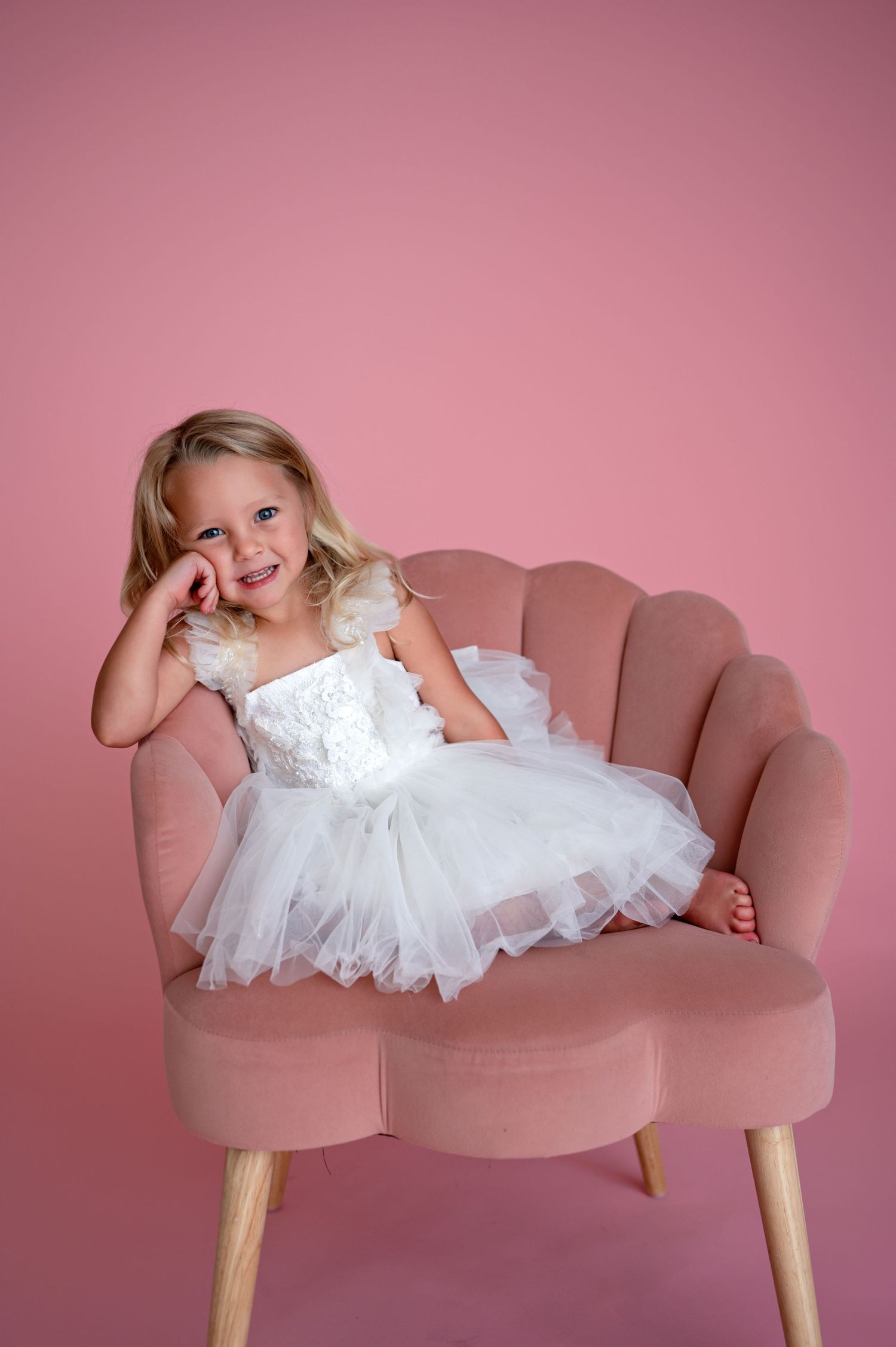 A child wearing a white tutu dress sits casually on a pink scalloped accent chair against a rosy pink background.
