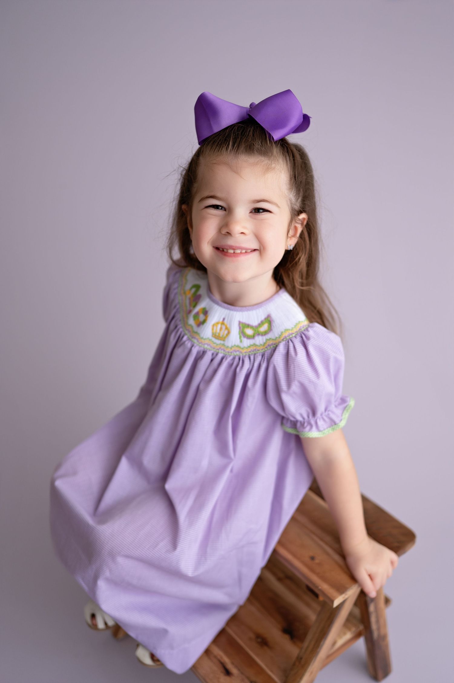 A young child in a purple dress smiles and poses playfully on a wooden chair against a gray backdrop.