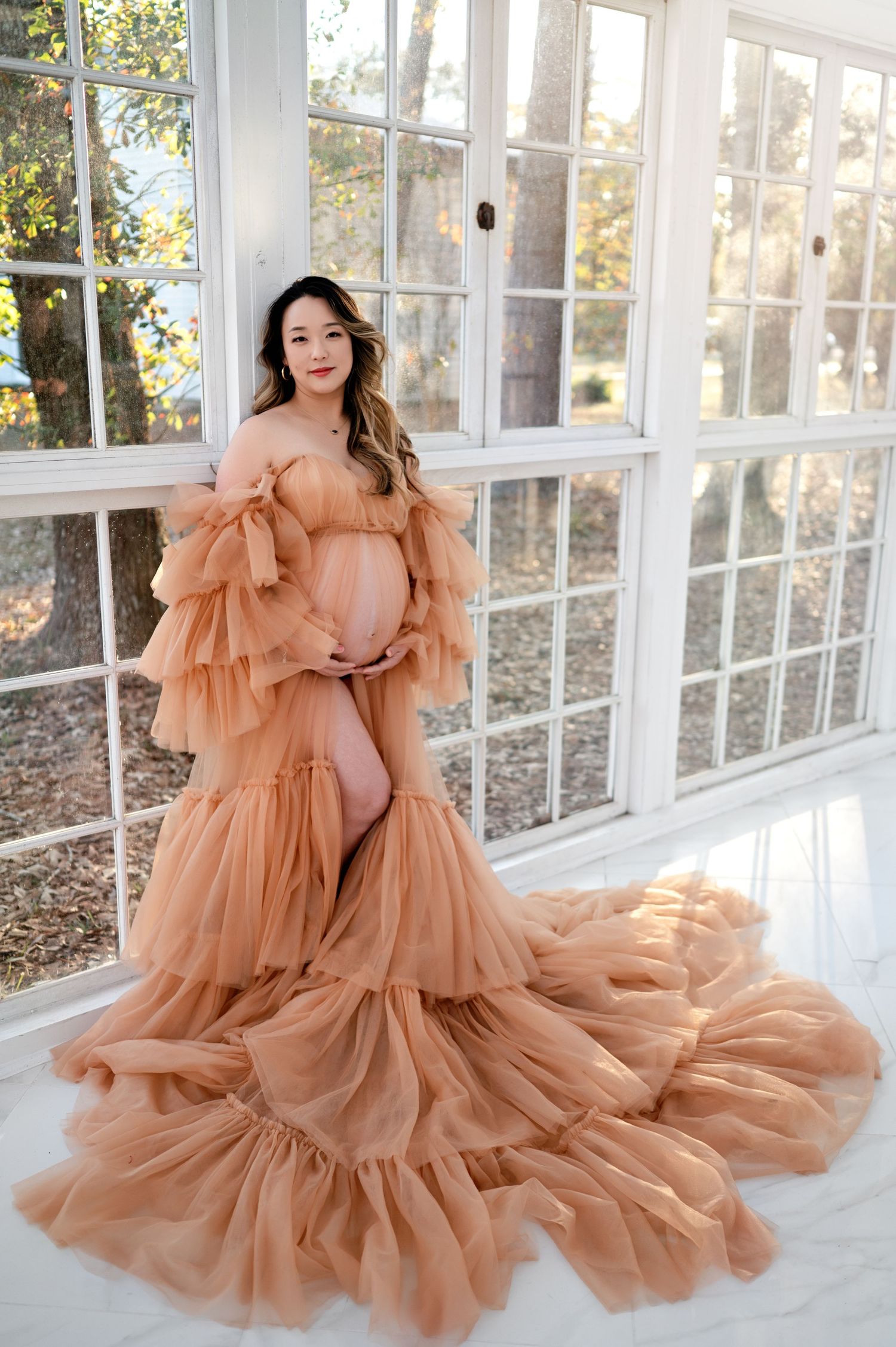 Ethereal peach-colored tulle maternity gown with ruffled tiers and flowing train in sunlit greenhouse setting.