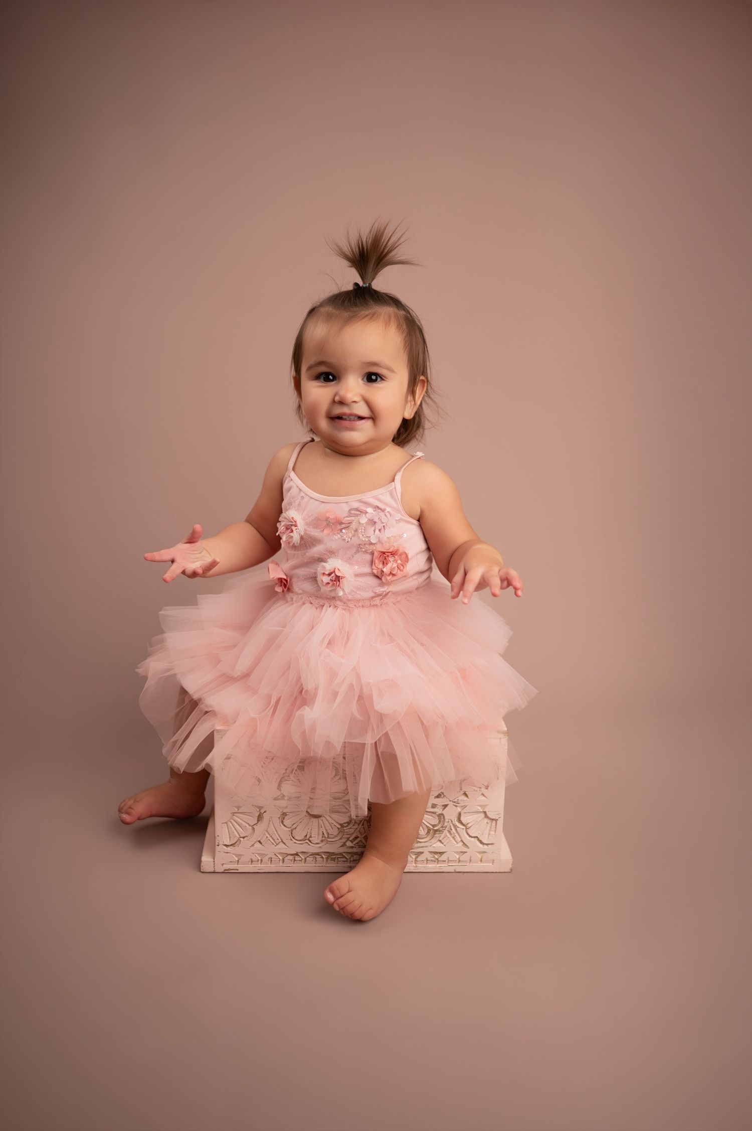 Baby wearing pink tulle dress sits on decorative box against neutral background in studio portrait.