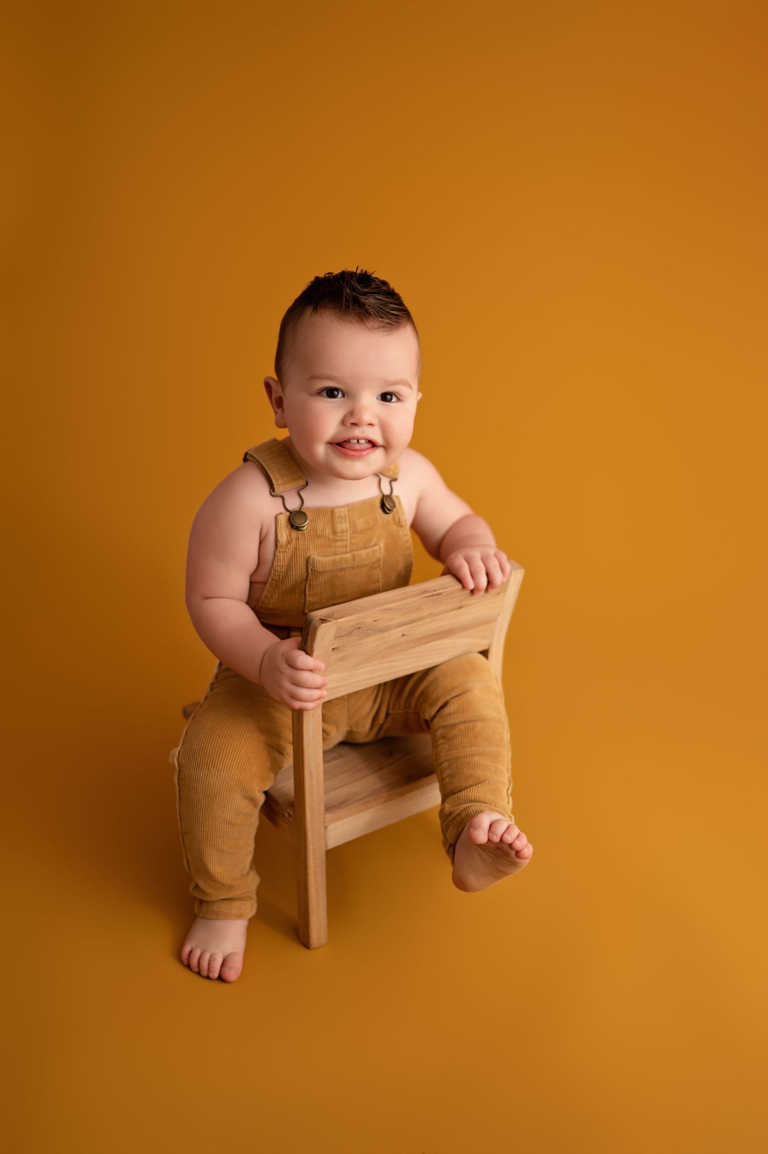 Baby in brown overalls sits and plays on a small wooden stool against an orange studio backdrop.