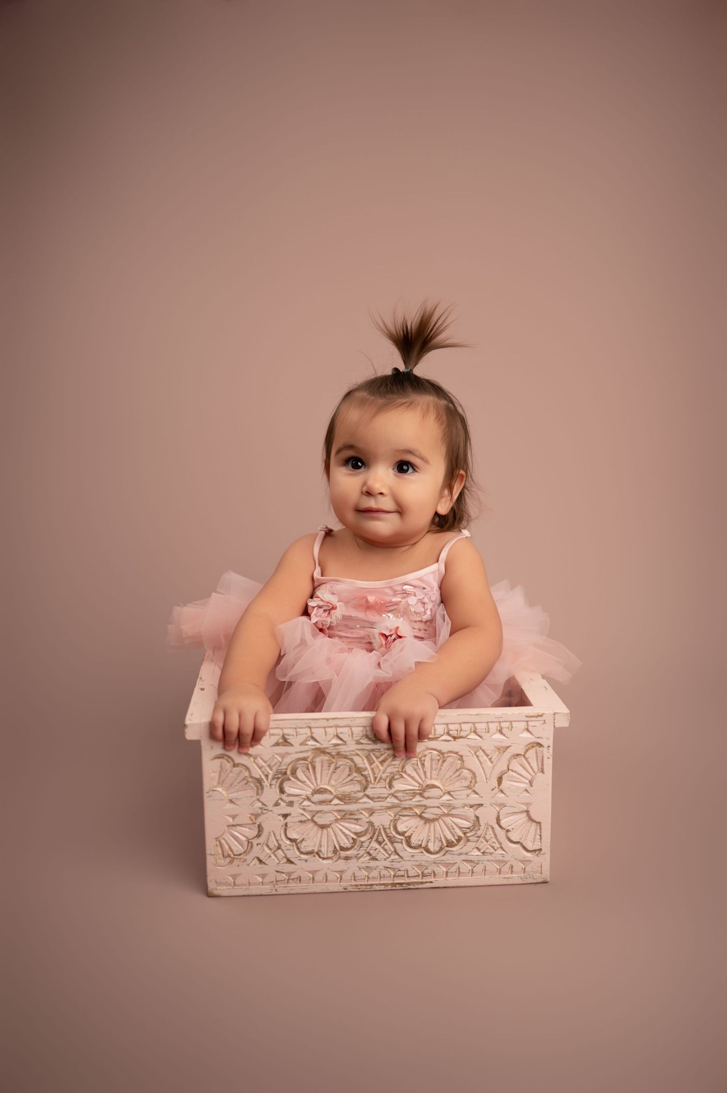 A baby in a pink dress sits in a decorative white box during a professional studio photo session.