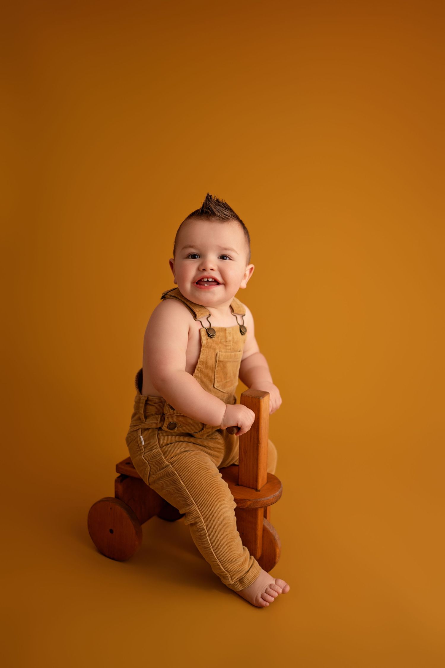 Baby in tan overalls sits against orange background with wooden rocking horse toy in studio portrait series.
