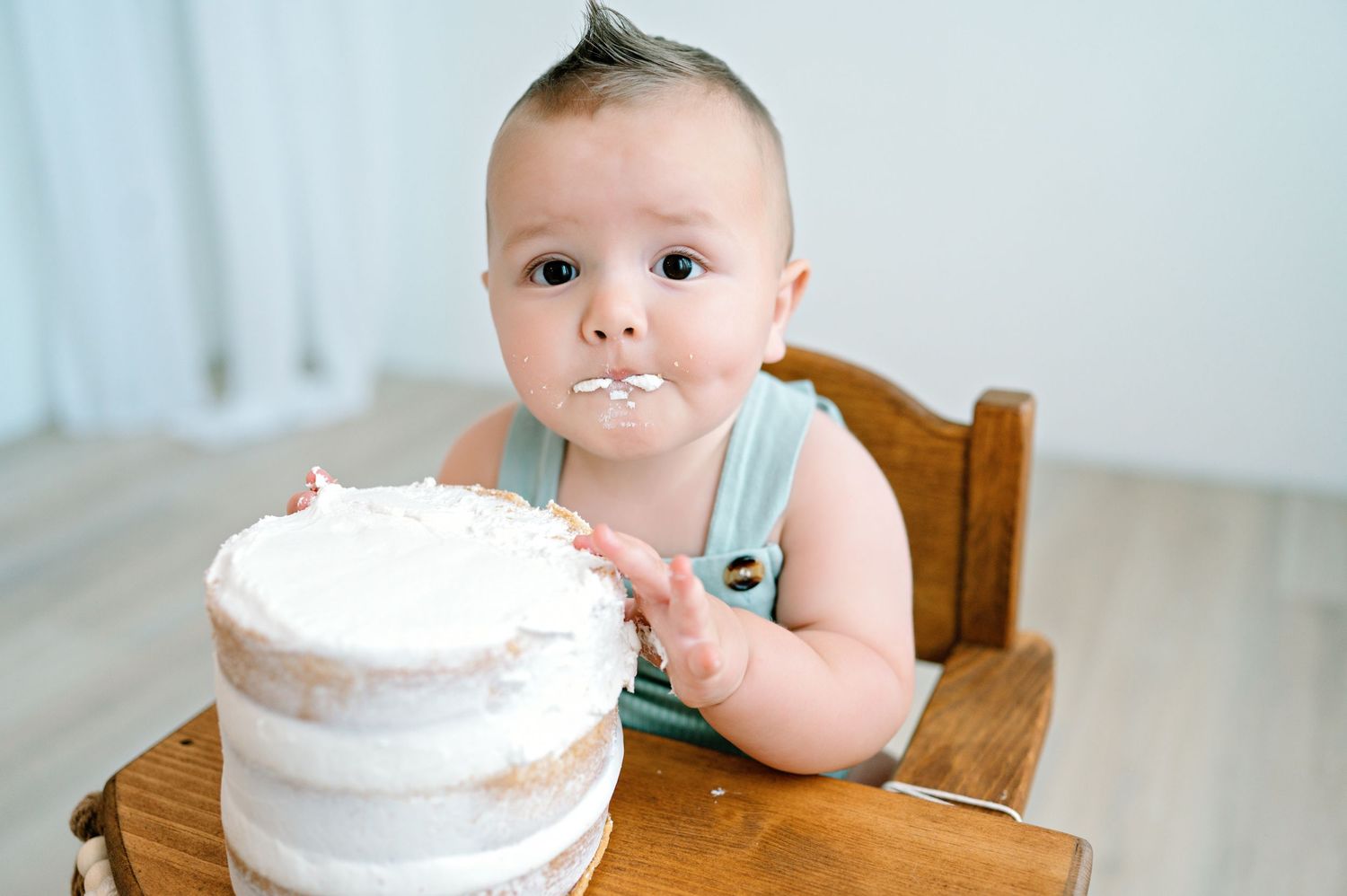 A baby sits in a wooden high chair enjoying a large white birthday cake with frosting smeared on their face.