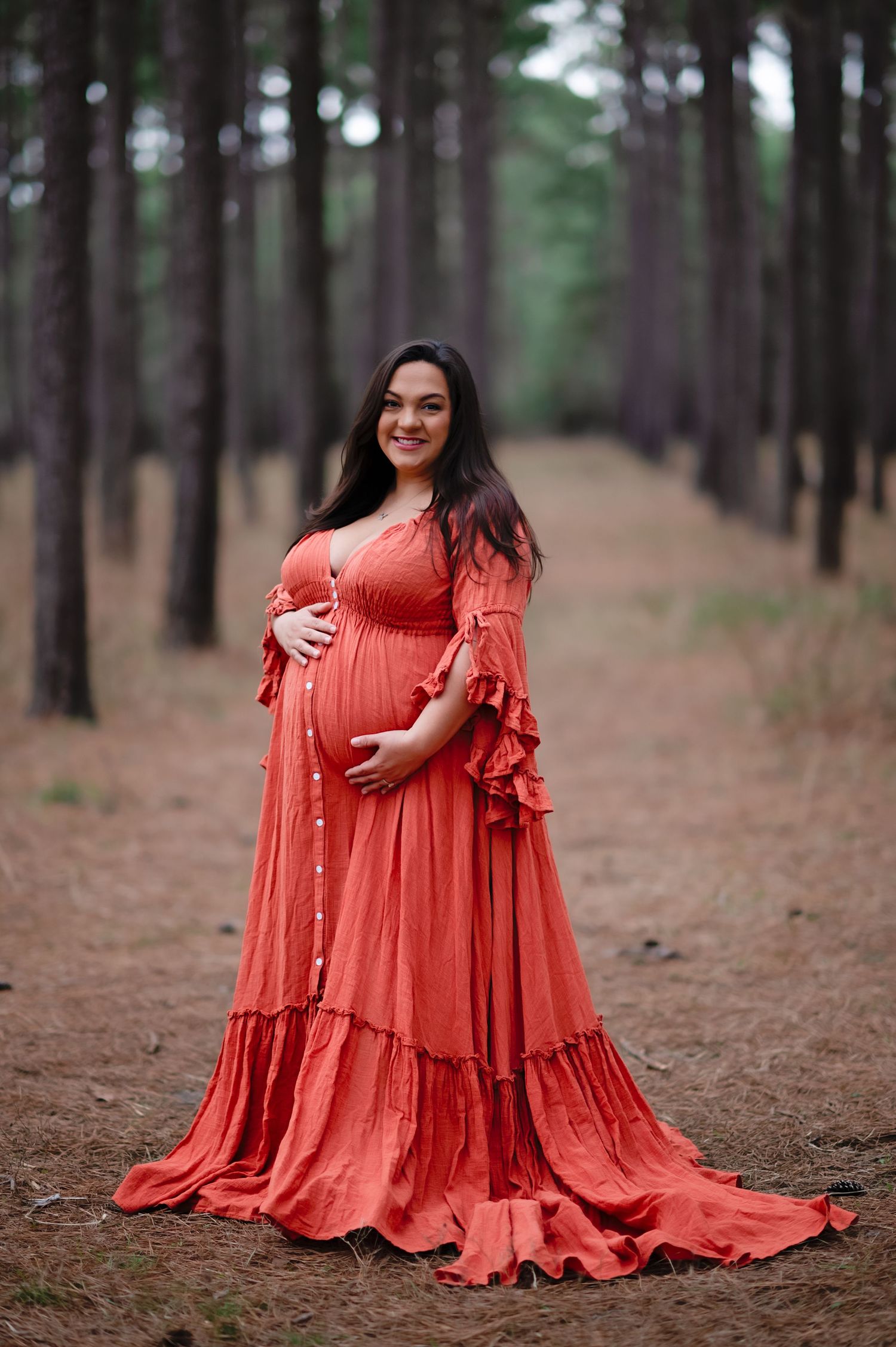Maternity photo in coral flowing dress amid pine forest setting.