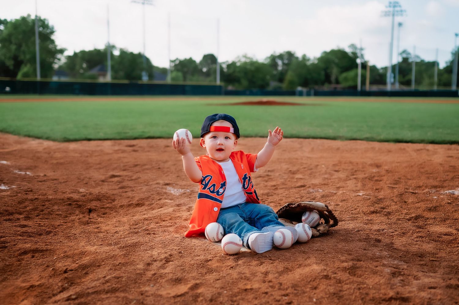 A small child in orange uniform sits on a baseball field dirt with arms raised in excitement.