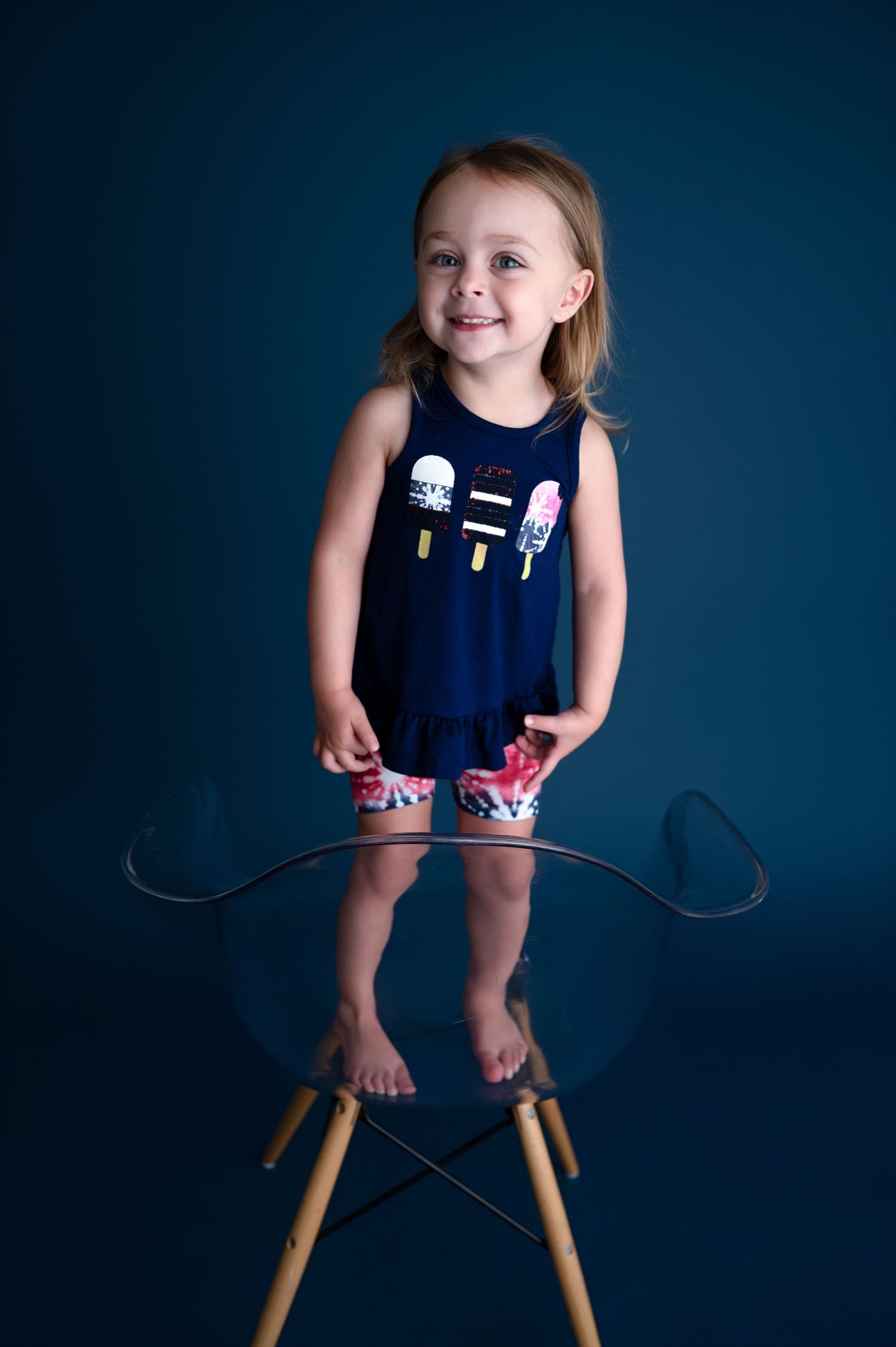 Happy child in navy ice cream print outfit standing on transparent modern chair.