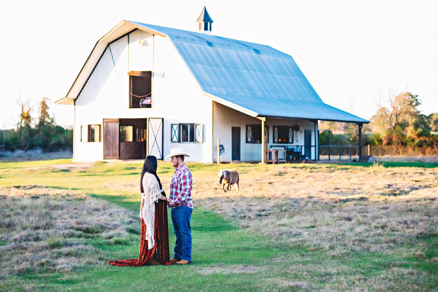 A couple stands together on their farmland with a white and blue barn in the background.