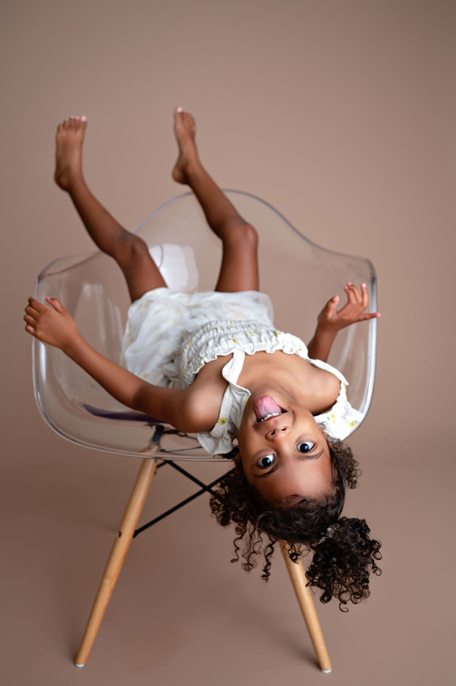 A child lays upside down in a transparent modern chair while wearing a white dress and having fun.