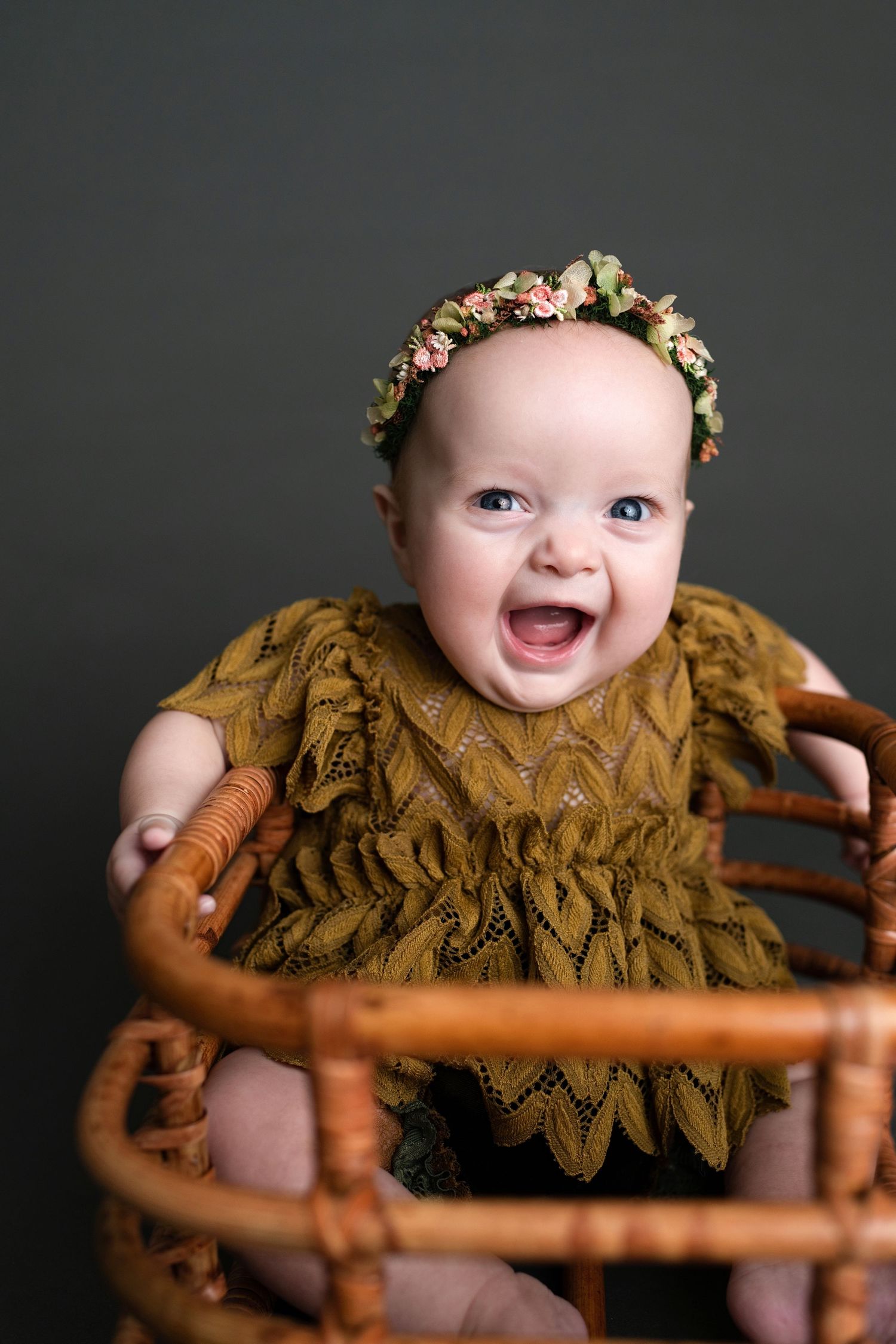 Baby wearing a floral crown and brown lace dress smiles while posing against a dark background in a photo series.