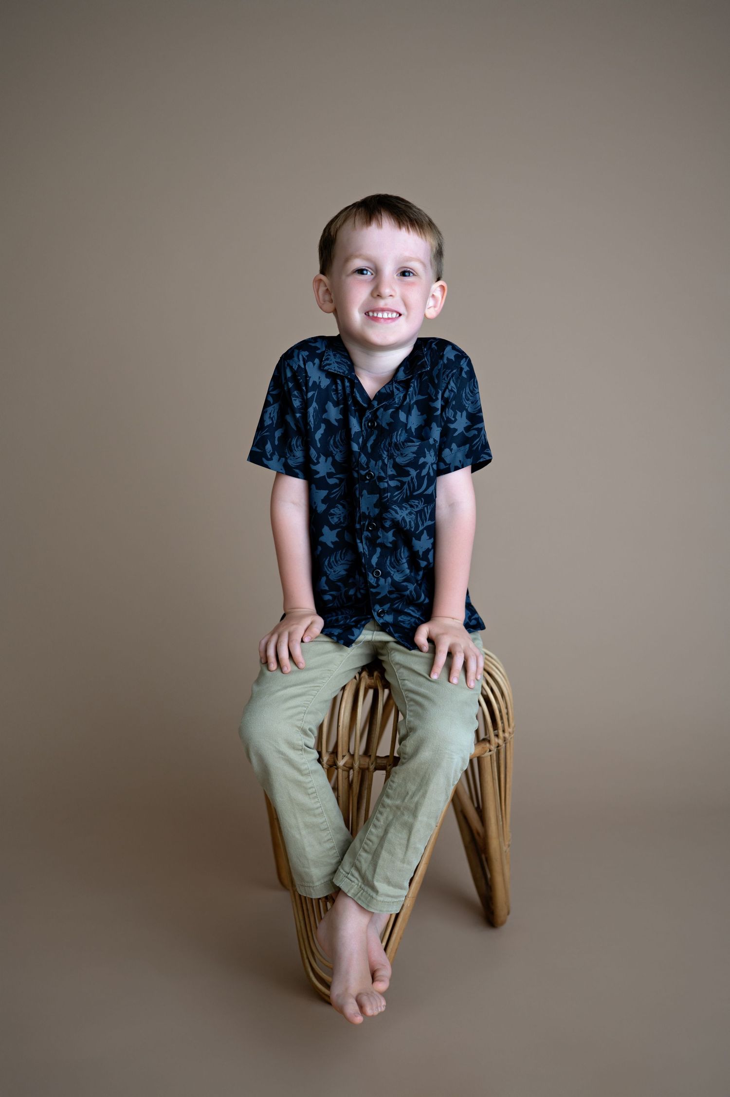 A young child wearing a navy floral shirt and khaki pants sits on a wooden stool against a neutral background.