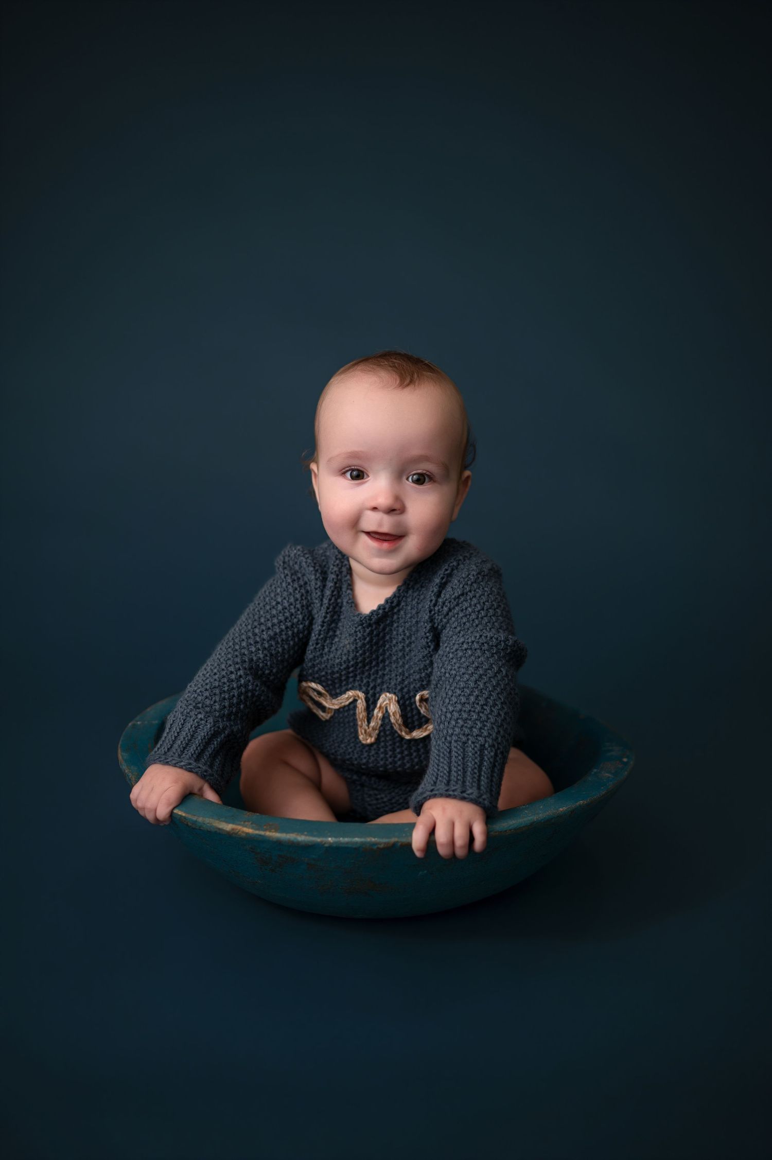 Baby wearing gray knit sweater sits and smiles while playing in a curved blue seat against dark background.