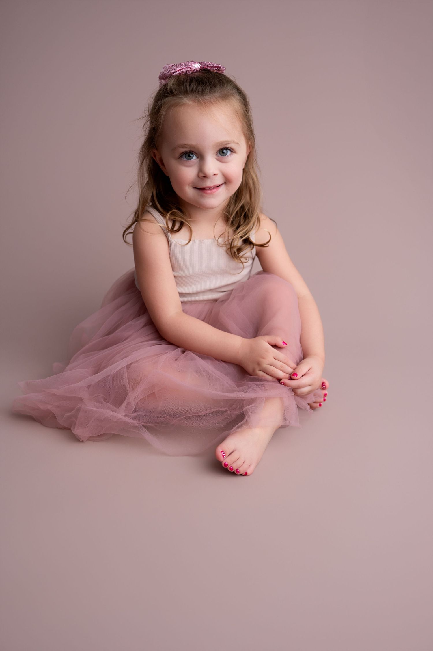 Child in pink tulle skirt and white top poses gracefully on pink studio backdrop.