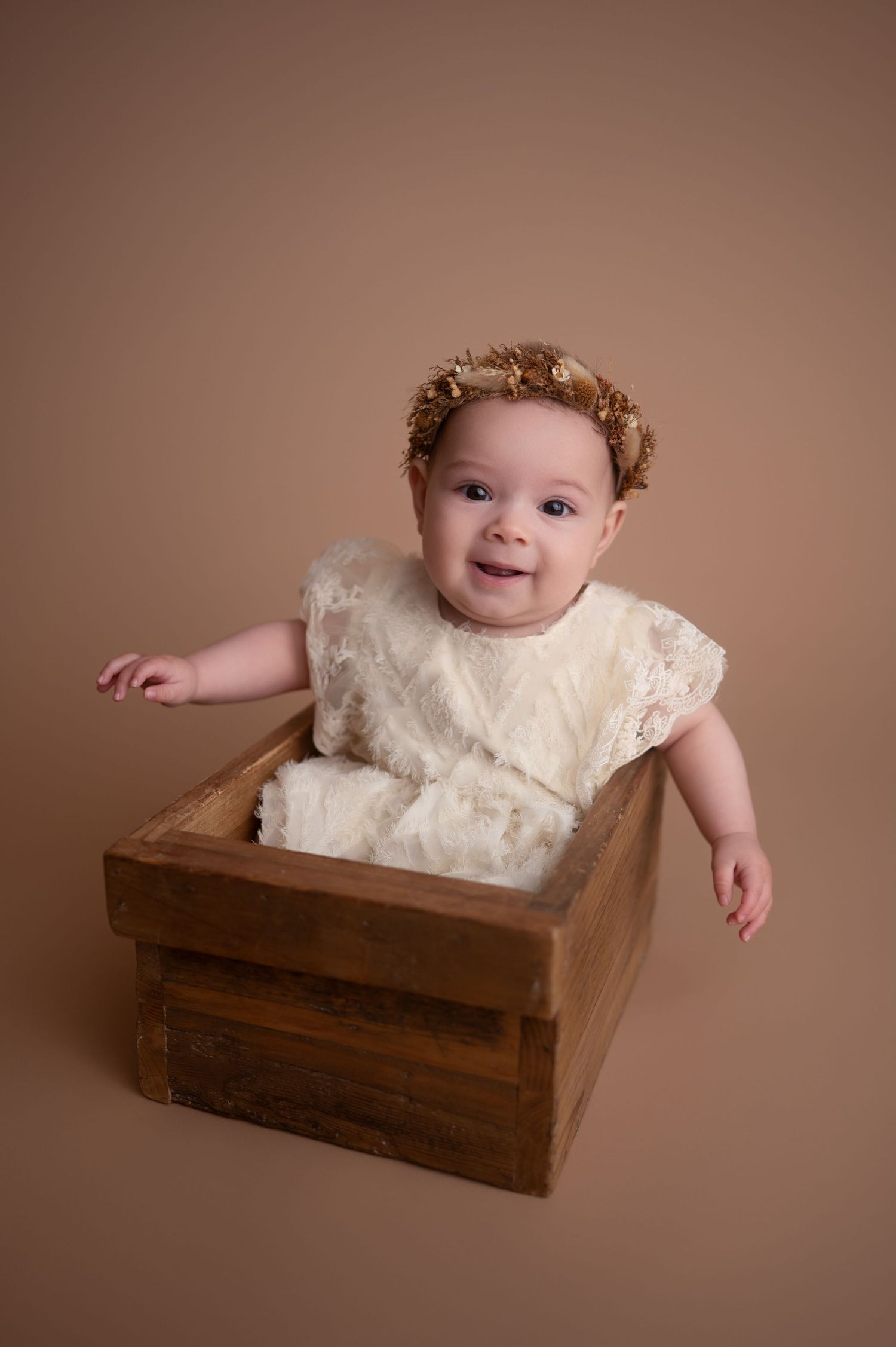 A baby wearing a cream lace dress sits in a rustic wooden crate during a professional photo session.