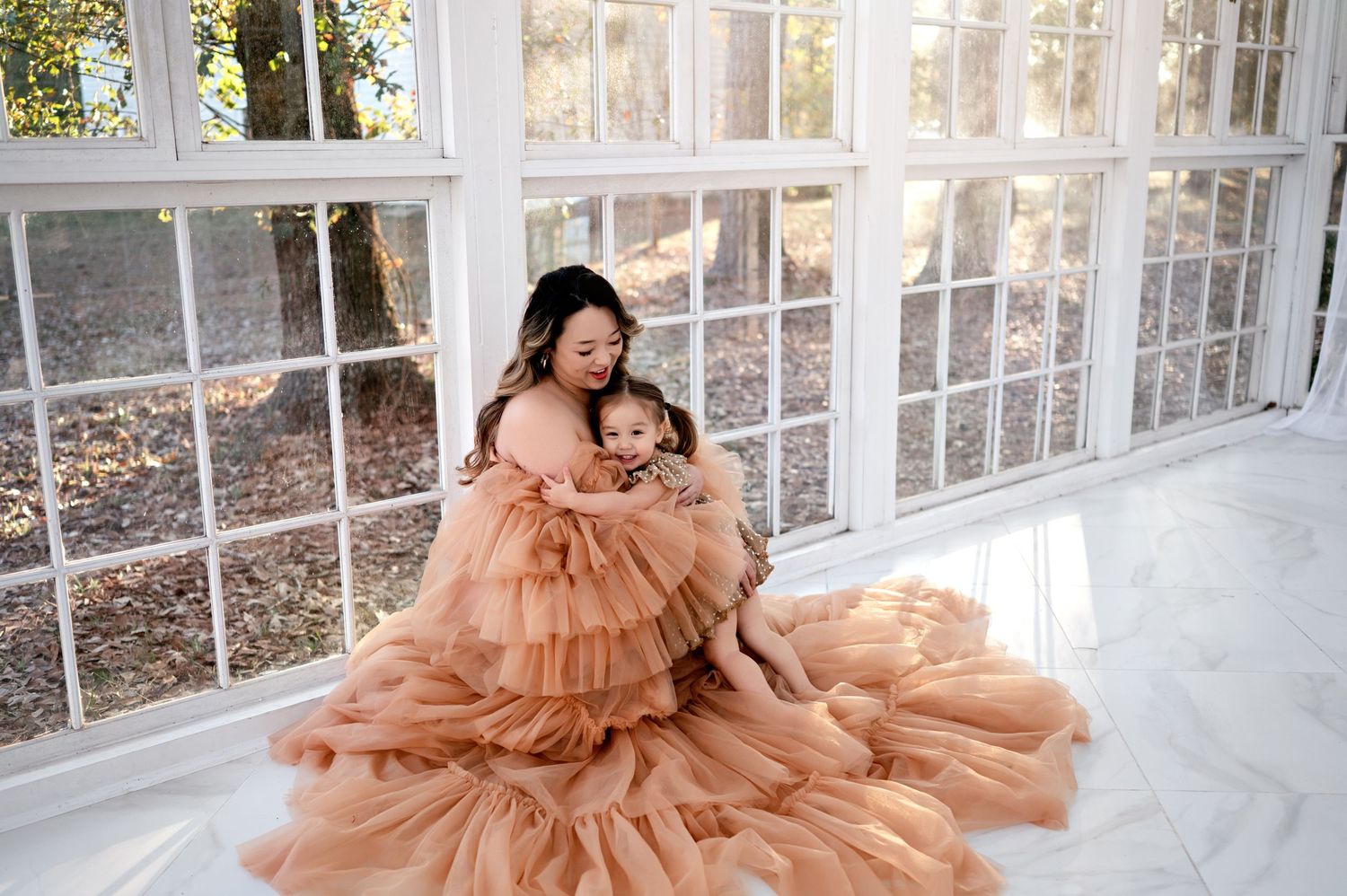 A person in a flowing peach tulle gown poses gracefully in a sunlit conservatory setting.