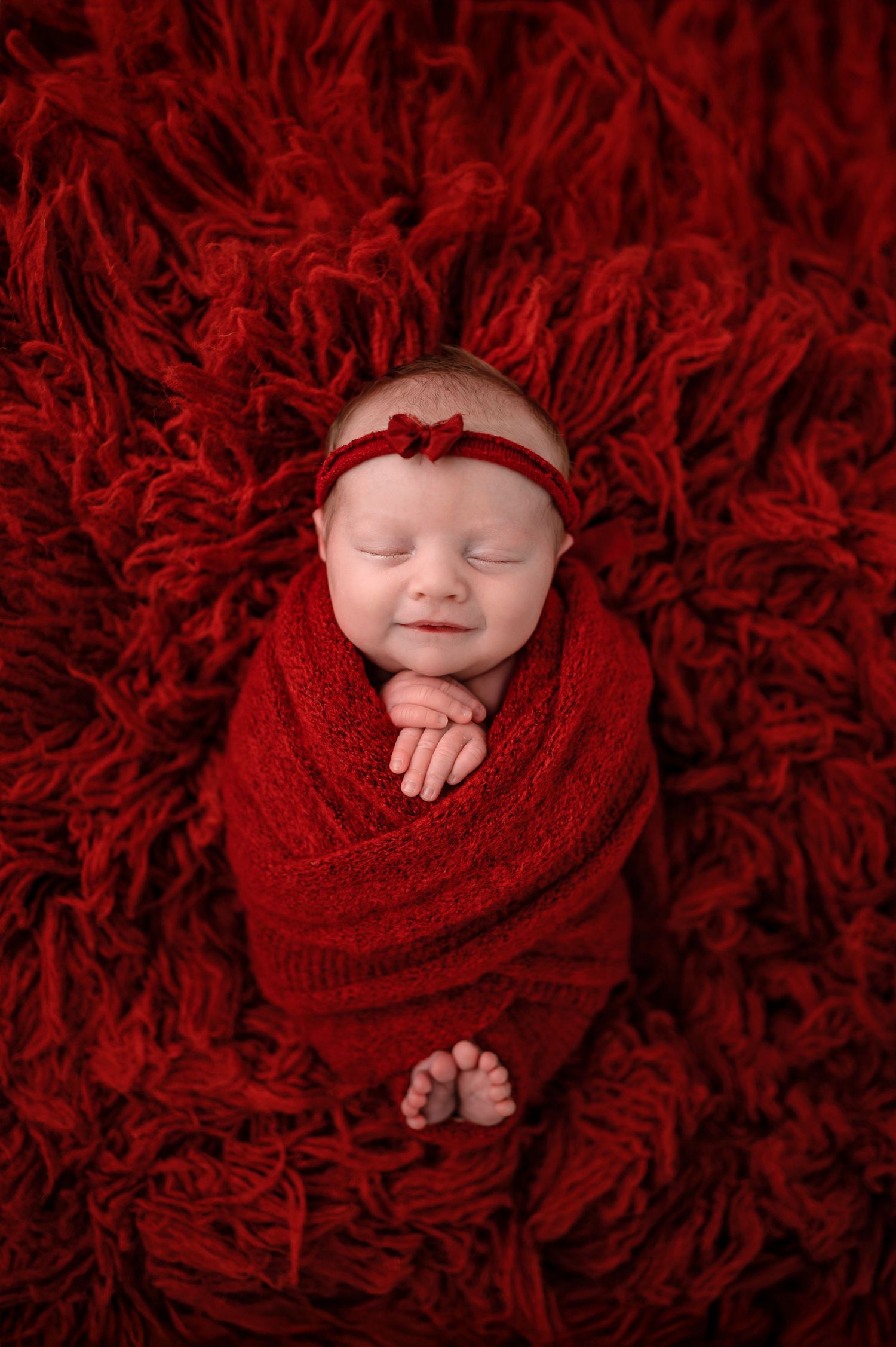 A sleeping baby swaddled in red fabric lies on a vibrant red textured blanket in a professional newborn photo shoot.