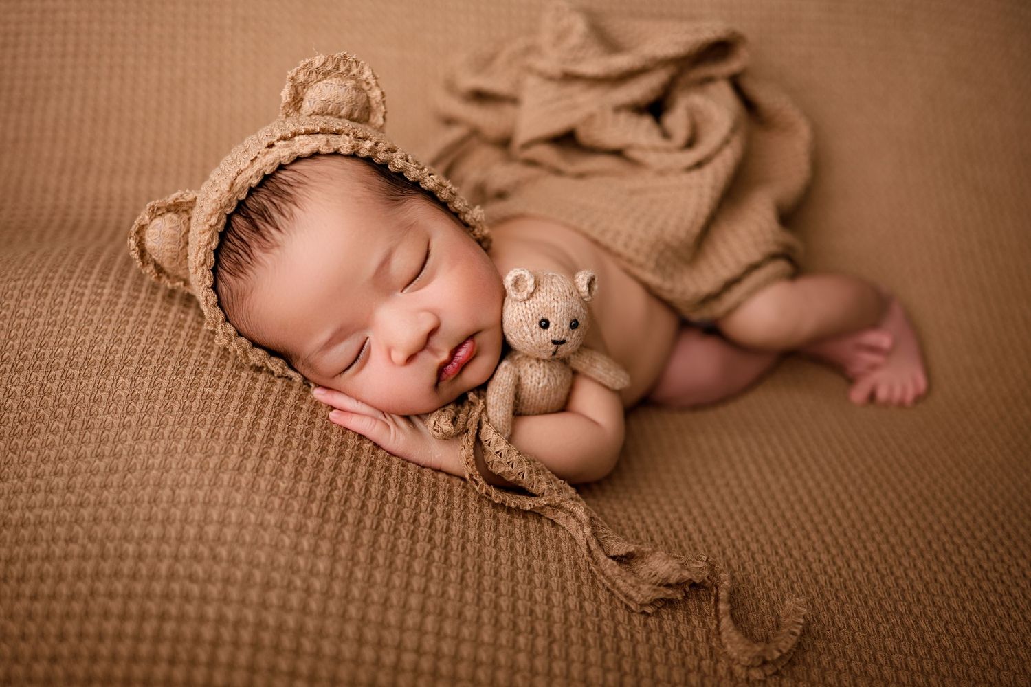 Newborn baby sleeping peacefully wearing a knit bear bonnet, cuddling a tiny teddy bear on a brown textured blanket.