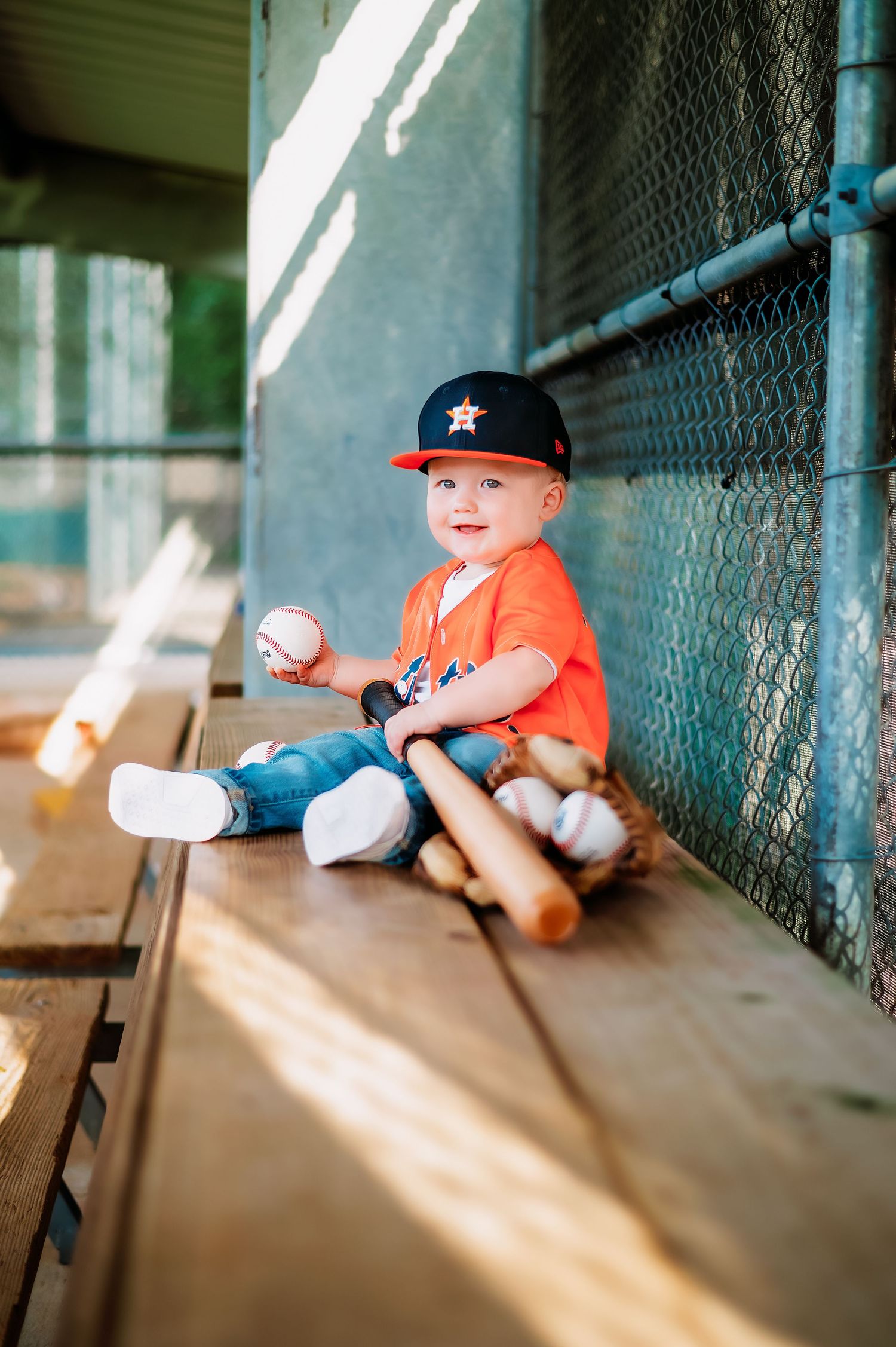 A young player in orange uniform sits in a dugout holding a baseball.