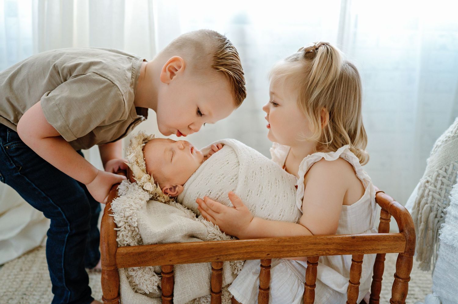 Two young children lean over a wooden crib to look at a baby in white, backlit by soft window light.