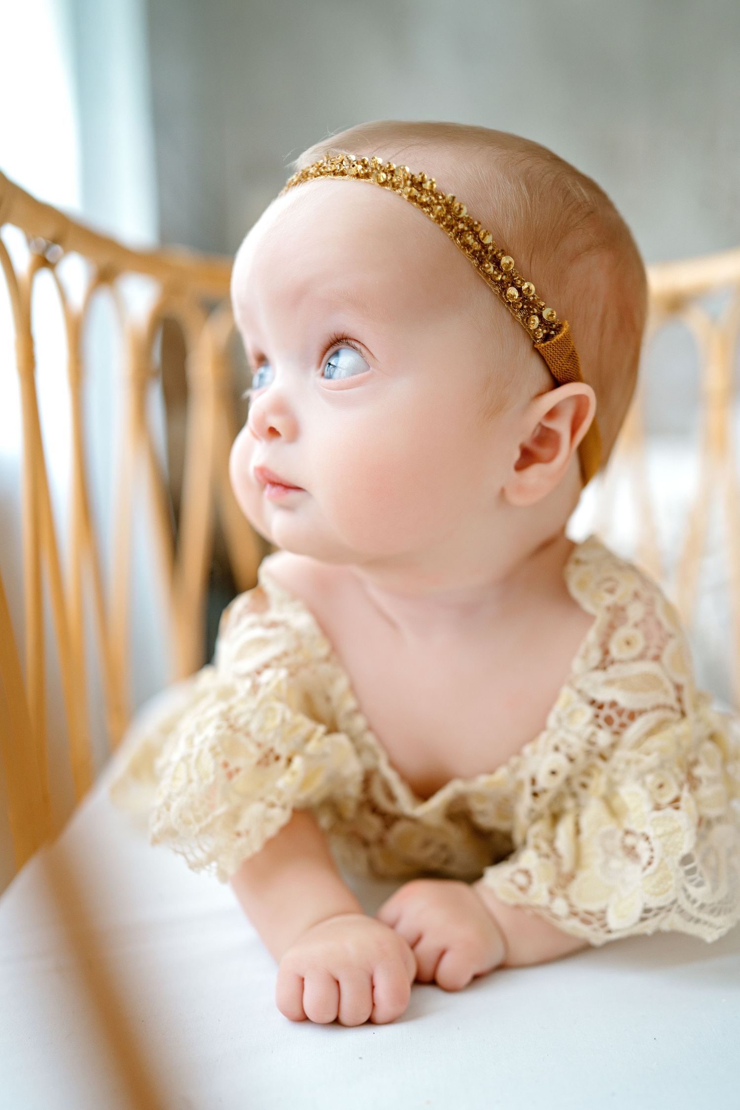 Baby wearing a delicate cream lace dress and gold glitter headband gazes upward while resting in a wicker chair.