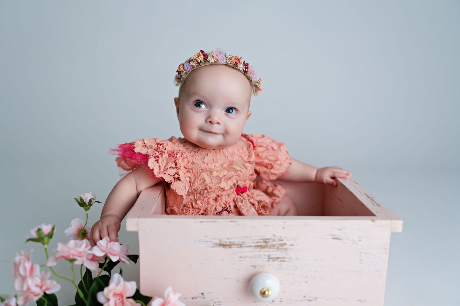 A baby in a peach lace dress and floral crown sits in a vintage pink drawer with flowers nearby.