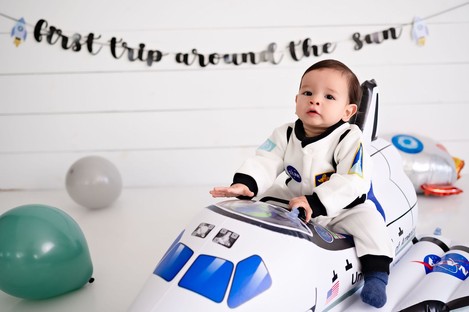 A small child dressed in a space-themed outfit sits in a toy rocket ship with colorful balloons in the background.