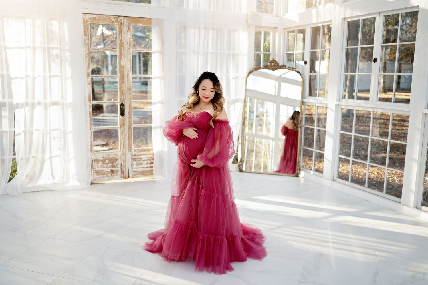 A woman in a flowing red tulle gown poses elegantly in a bright white room with large windows.