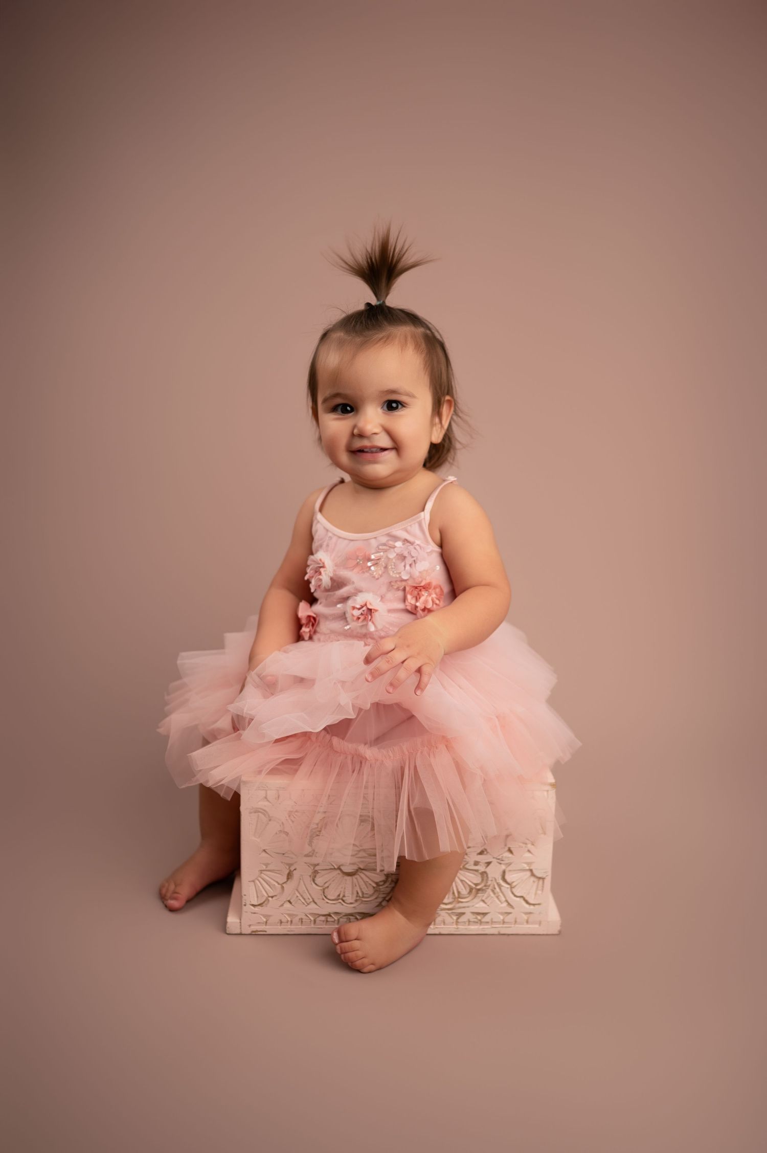 Baby wearing pink tulle dress sits on white box against mauve backdrop in professional portrait session.