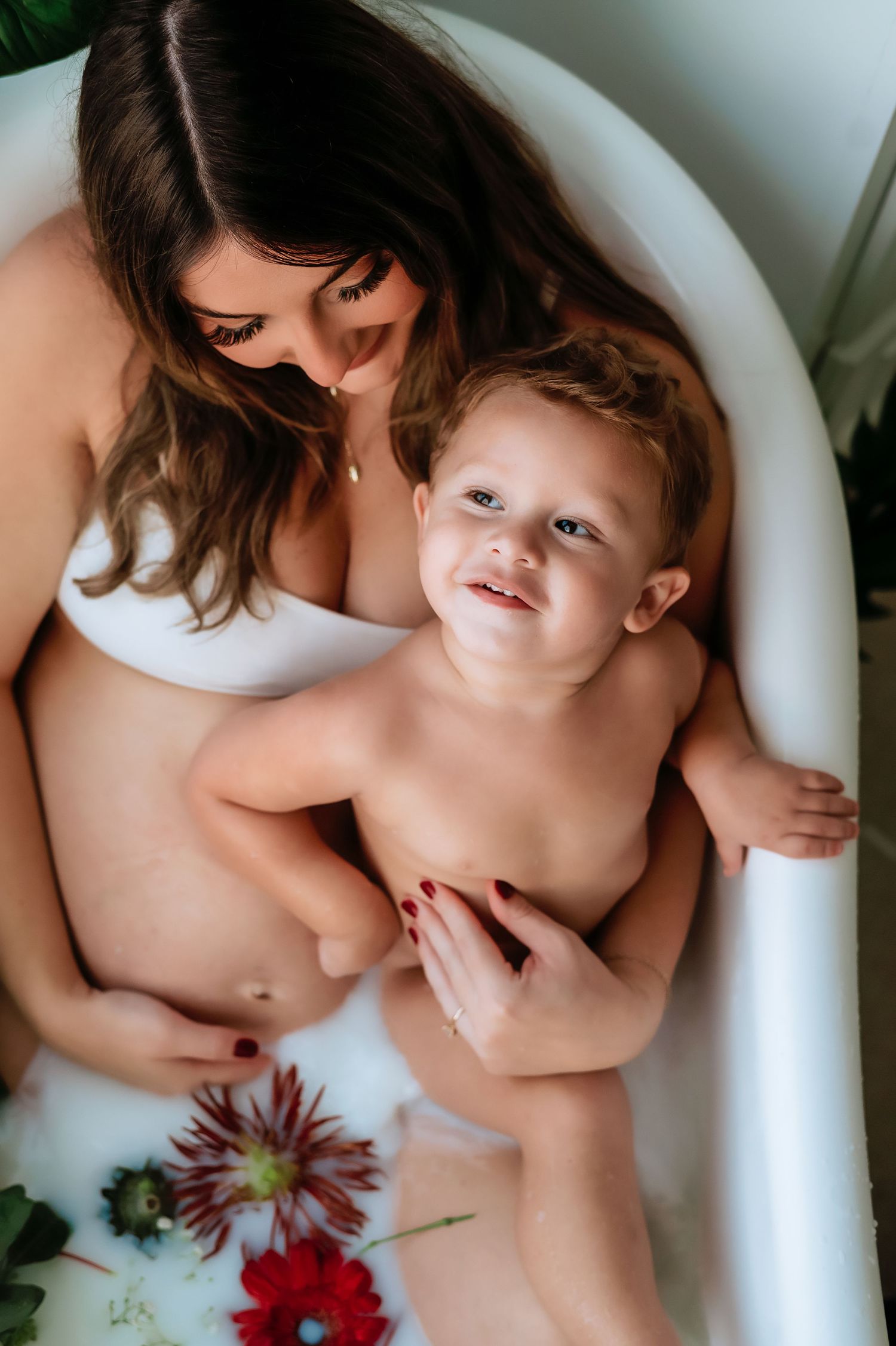 A tender maternity milk bath portrait with bright flowers and a joyful toddler.