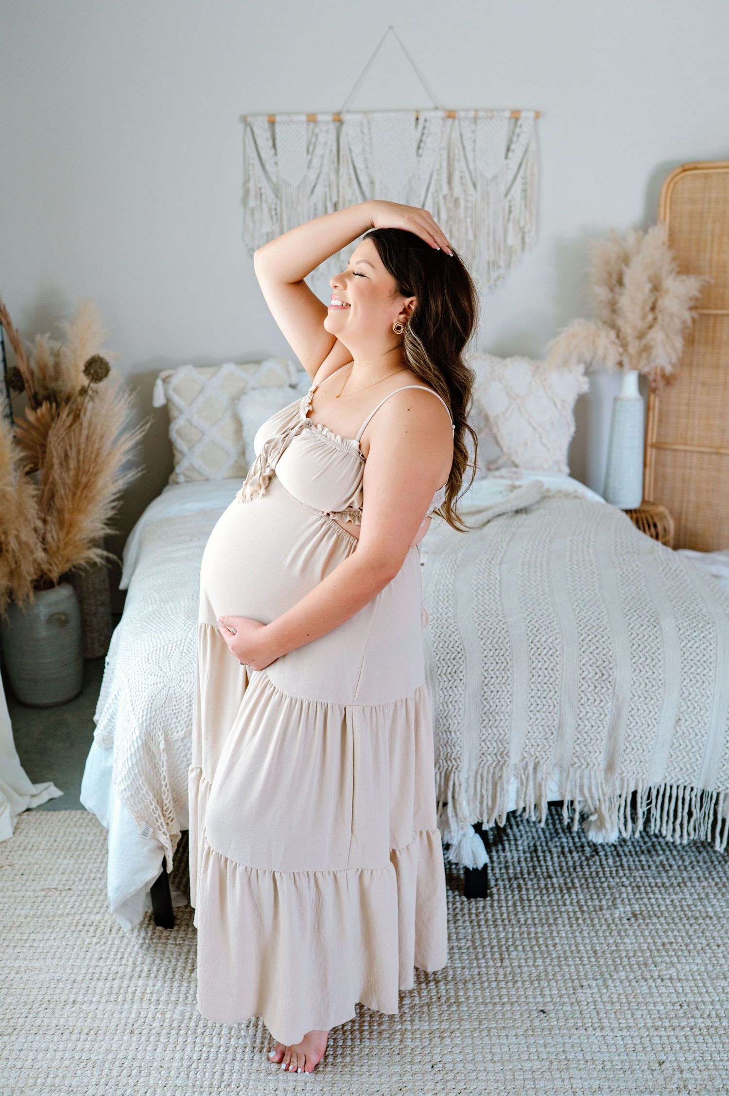 A pregnant woman in a flowing beige maxi dress stands in a cozy boho-style bedroom with dried pampas grass decor.