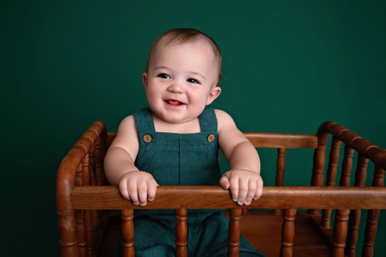 Infant in teal overalls smiles while standing in wooden crib against emerald backdrop.