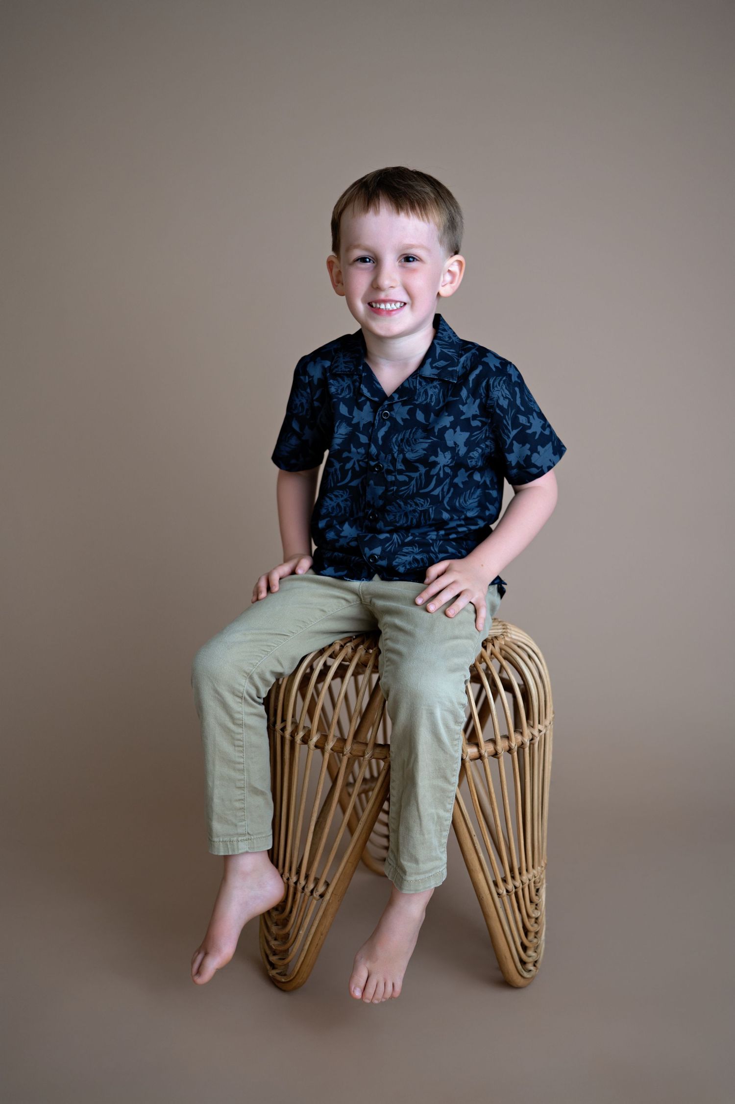 Portrait of child wearing navy patterned shirt and khaki pants sitting on rattan stool against beige backdrop.