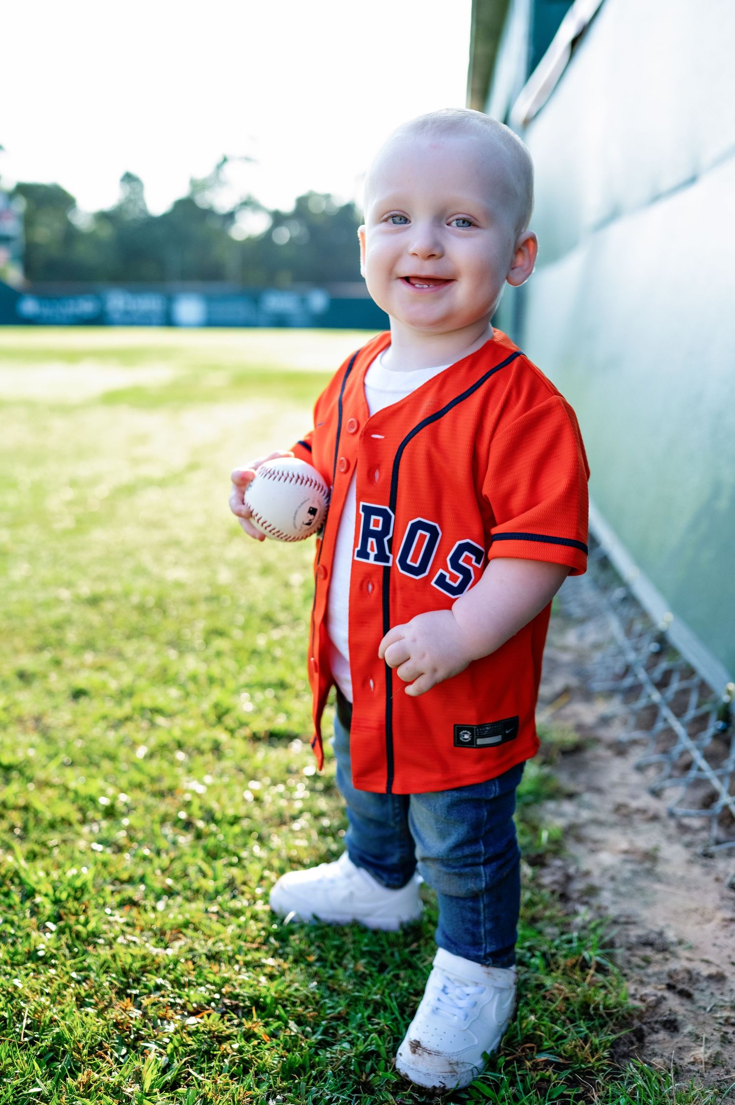 A baby wearing an orange baseball jersey and jeans plays with a baseball while standing on grass near a dugout.