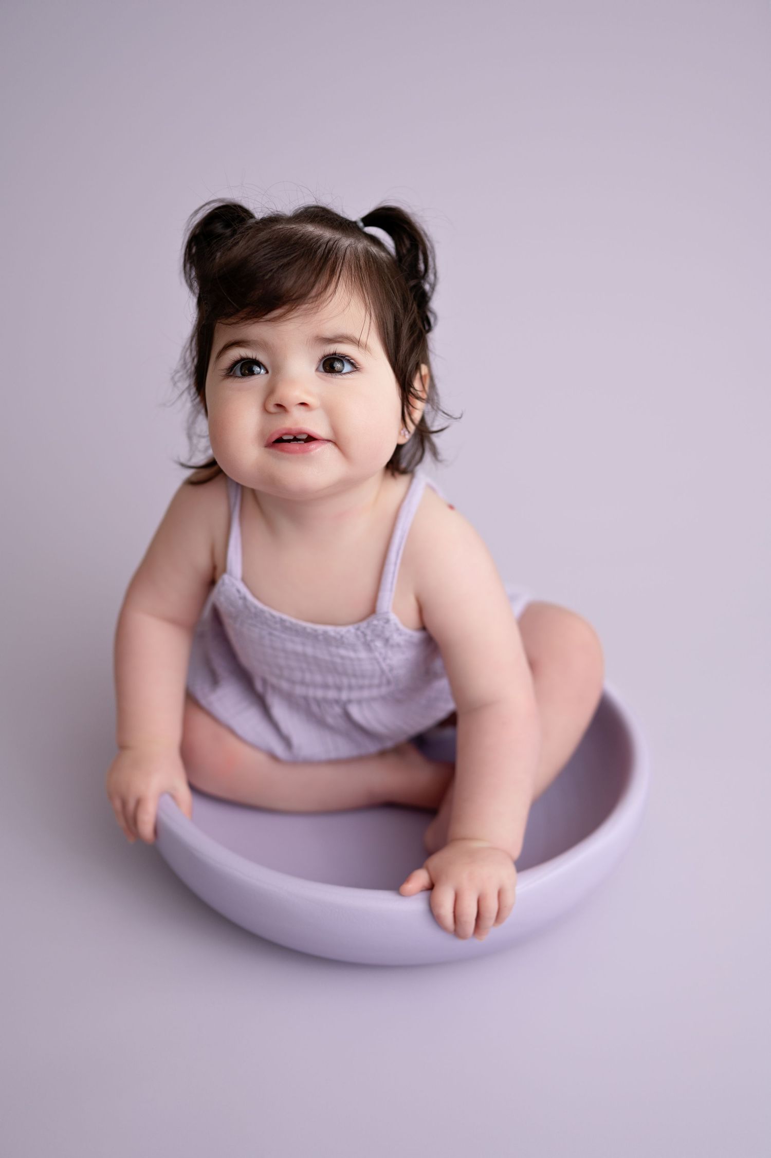 A baby in a lavender outfit sits in a curved bowl against a light purple background.