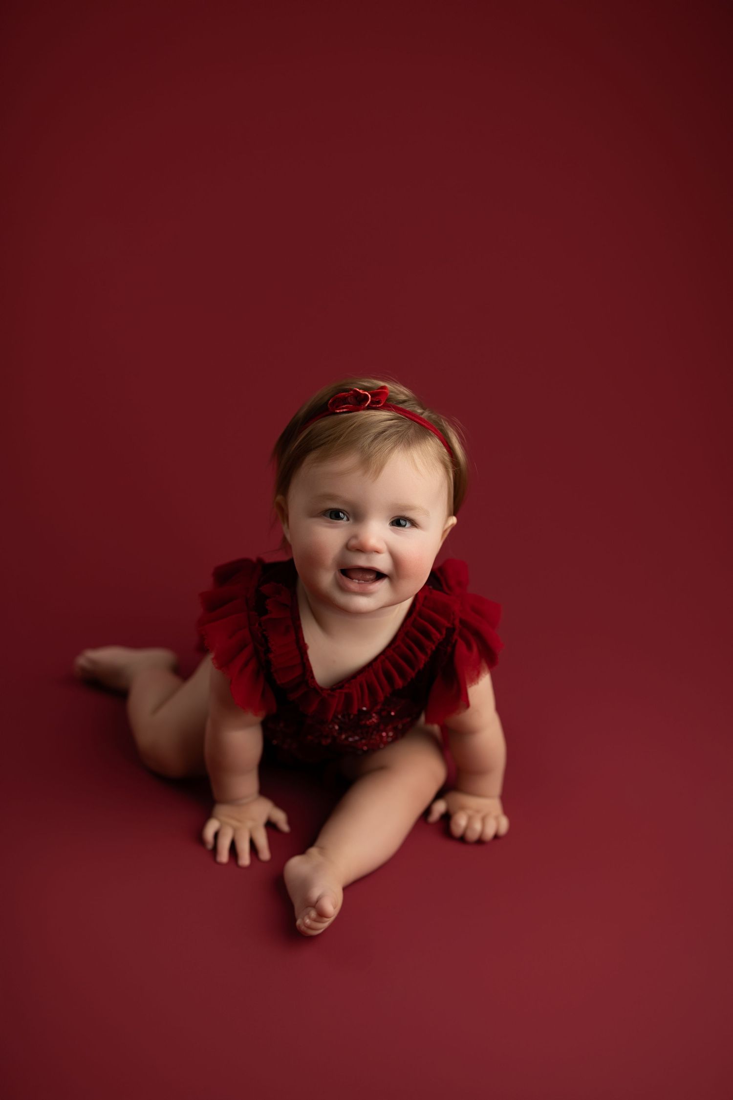 Baby in red dress crawls on burgundy backdrop during professional photo session.