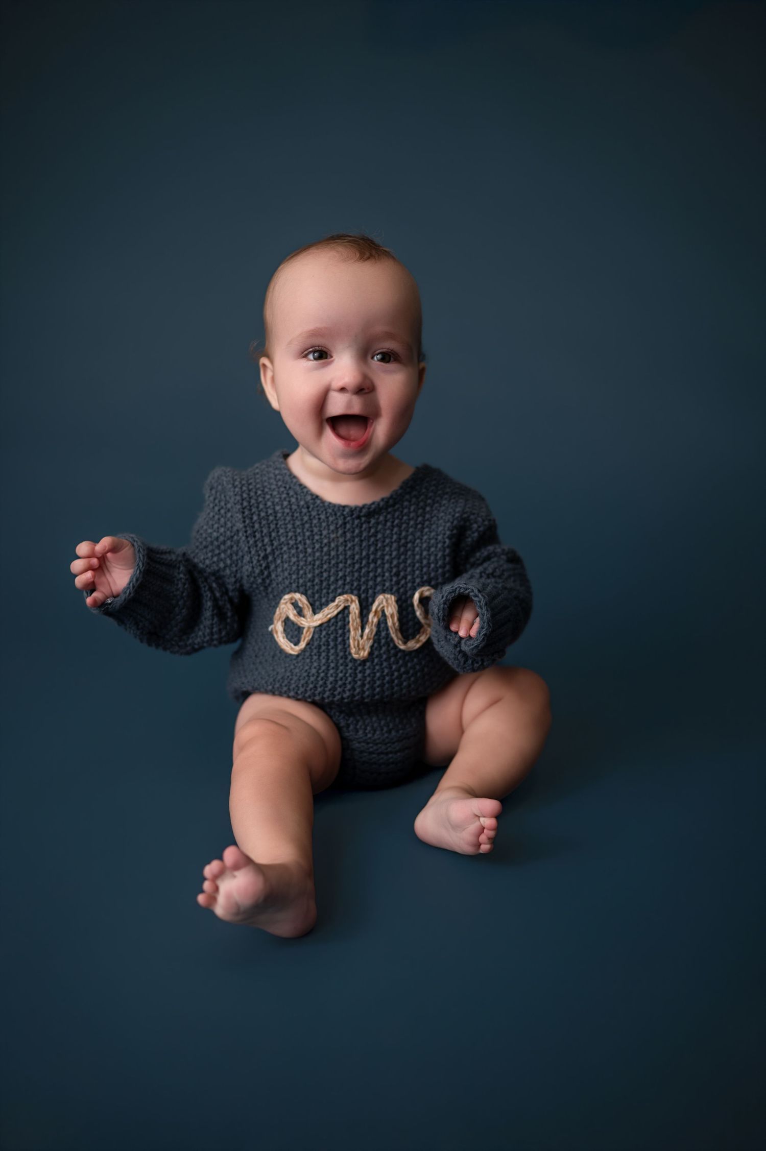 Baby wearing navy knit sweater lies on dark background in professional studio portrait series showing joyful expressions.