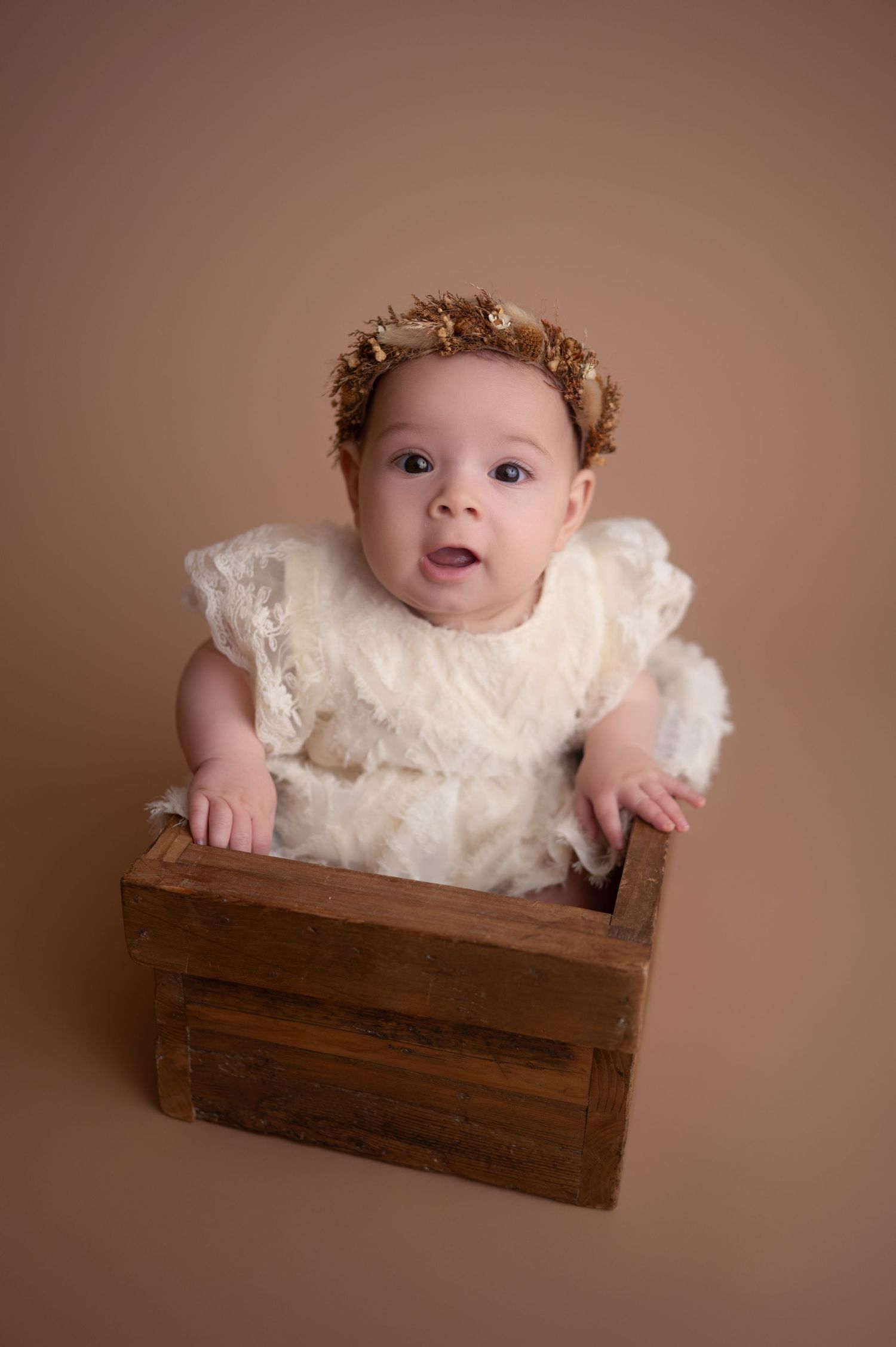 A baby in a white lace dress poses inside a rustic wooden crate against a brown backdrop in a studio setting.