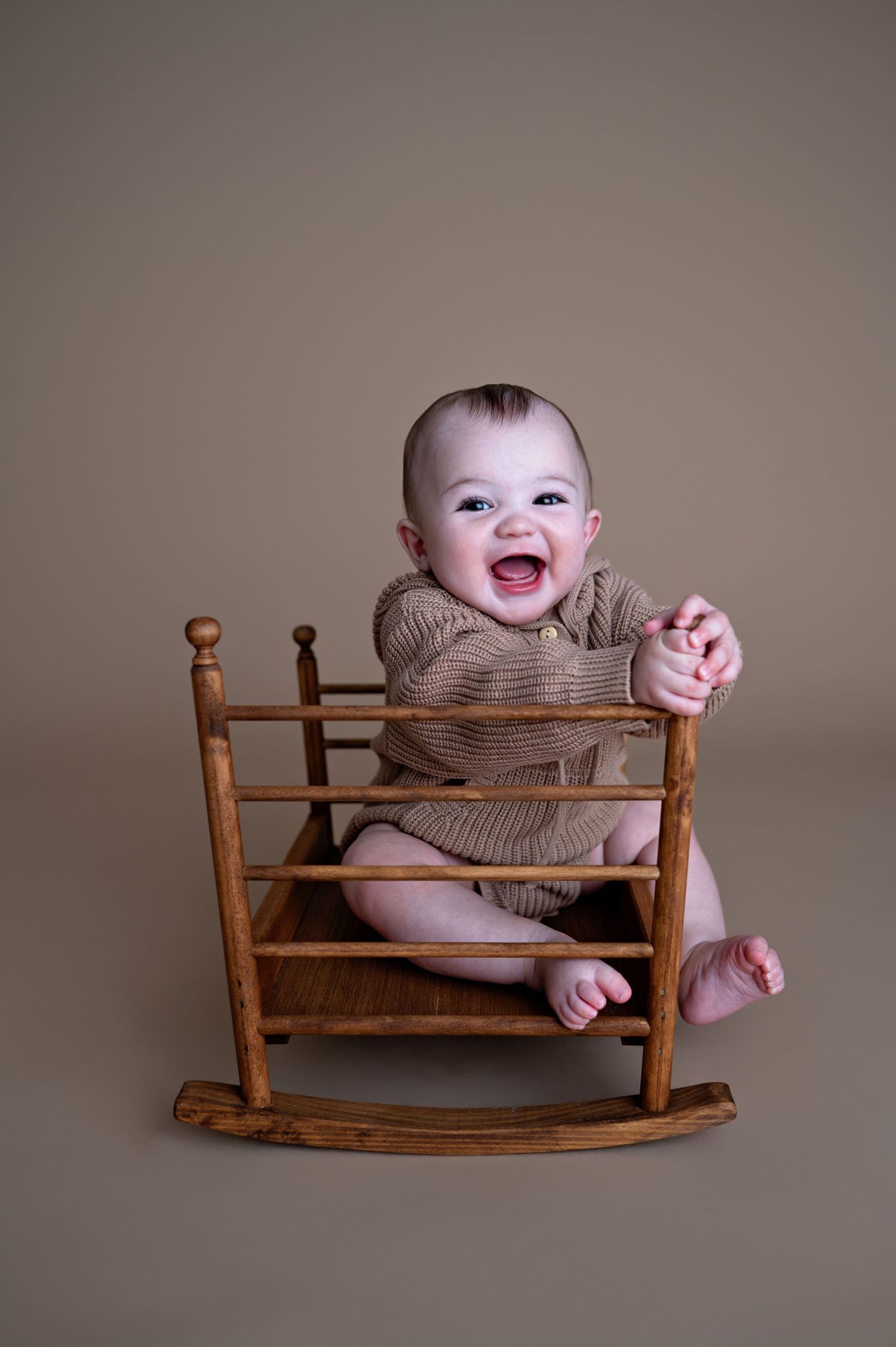 A joyful baby sitting in a wooden rocking chair against a neutral backdrop.