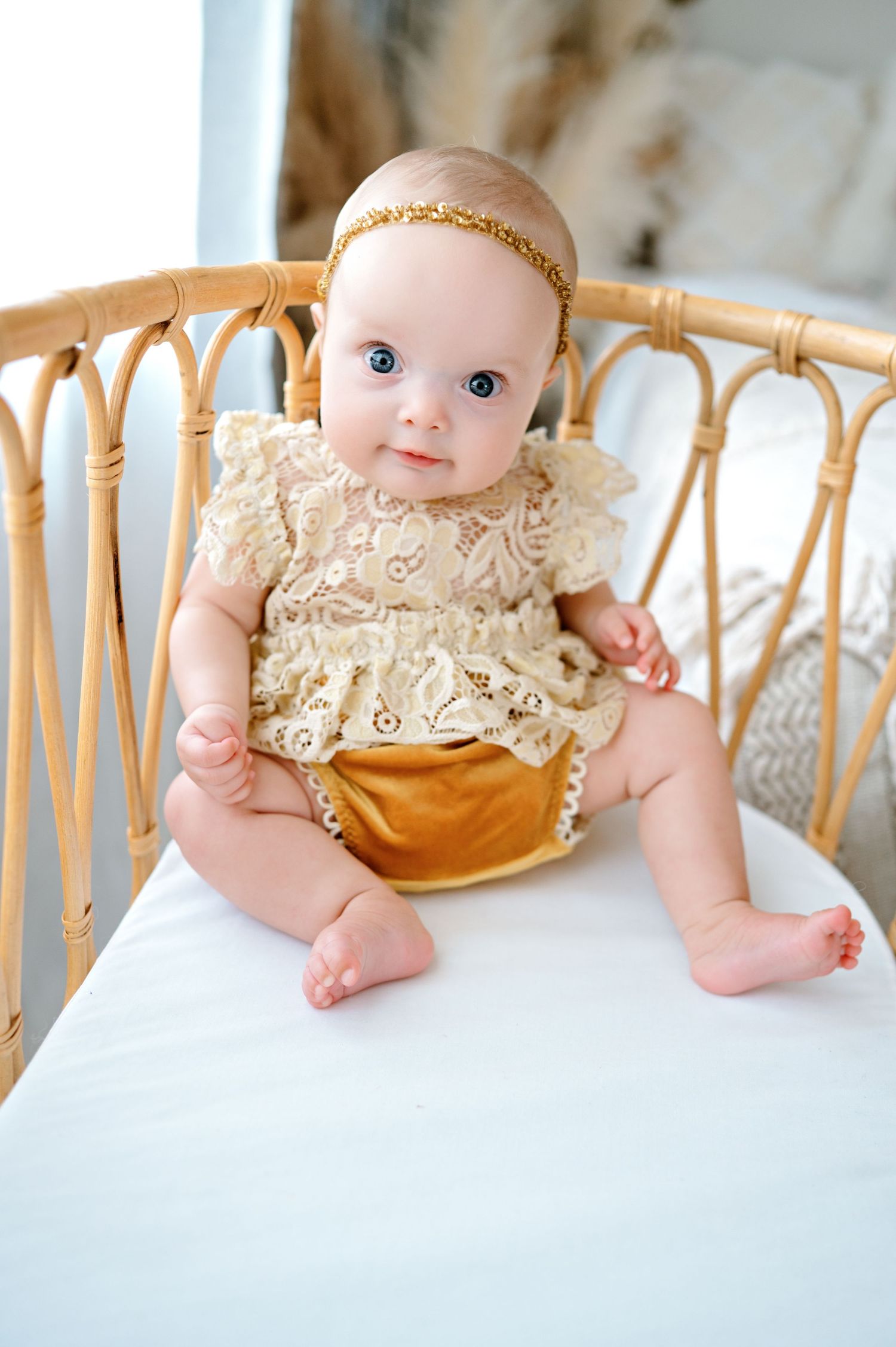 Baby wearing gold lace dress and headband lying on light colored furniture.