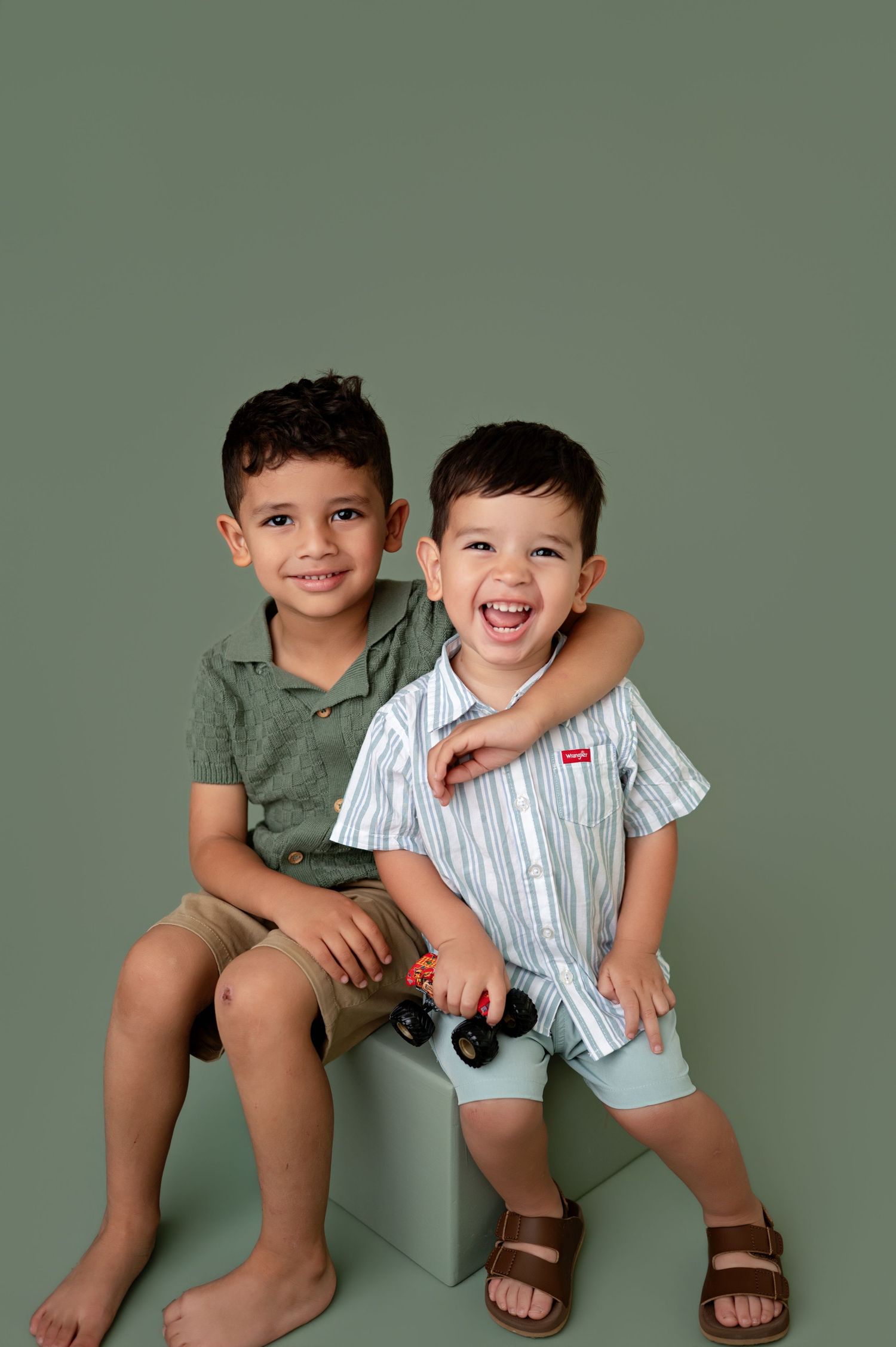 Two young brothers smiling and posing together on a white cube against a sage green background.