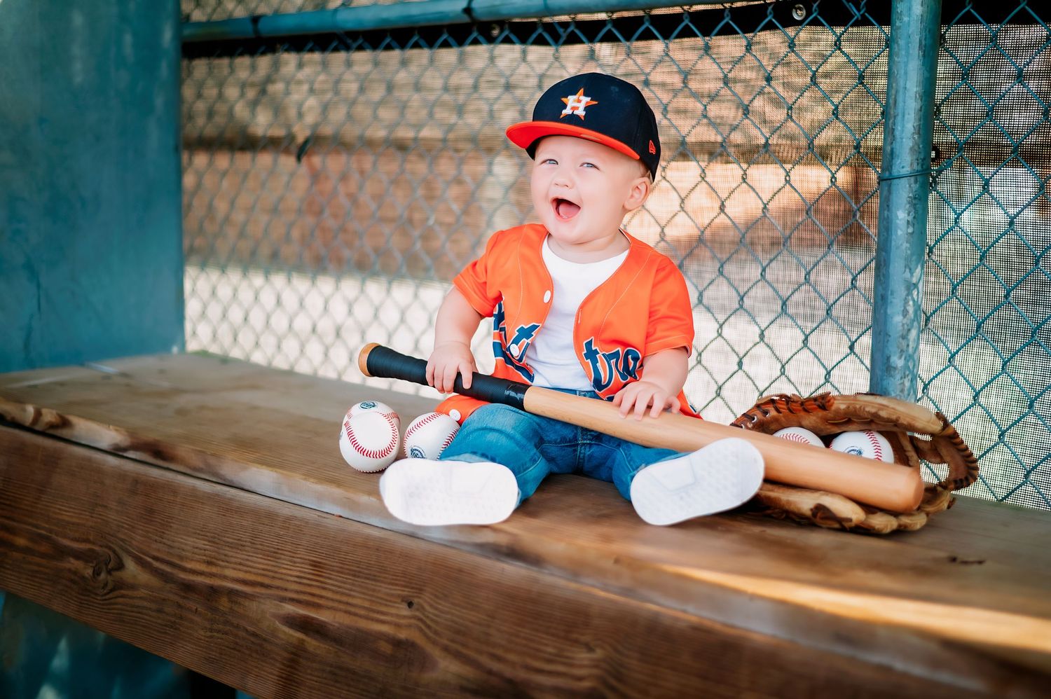 A small baseball player sits in a dugout with baseball and glove wearing an orange jersey.