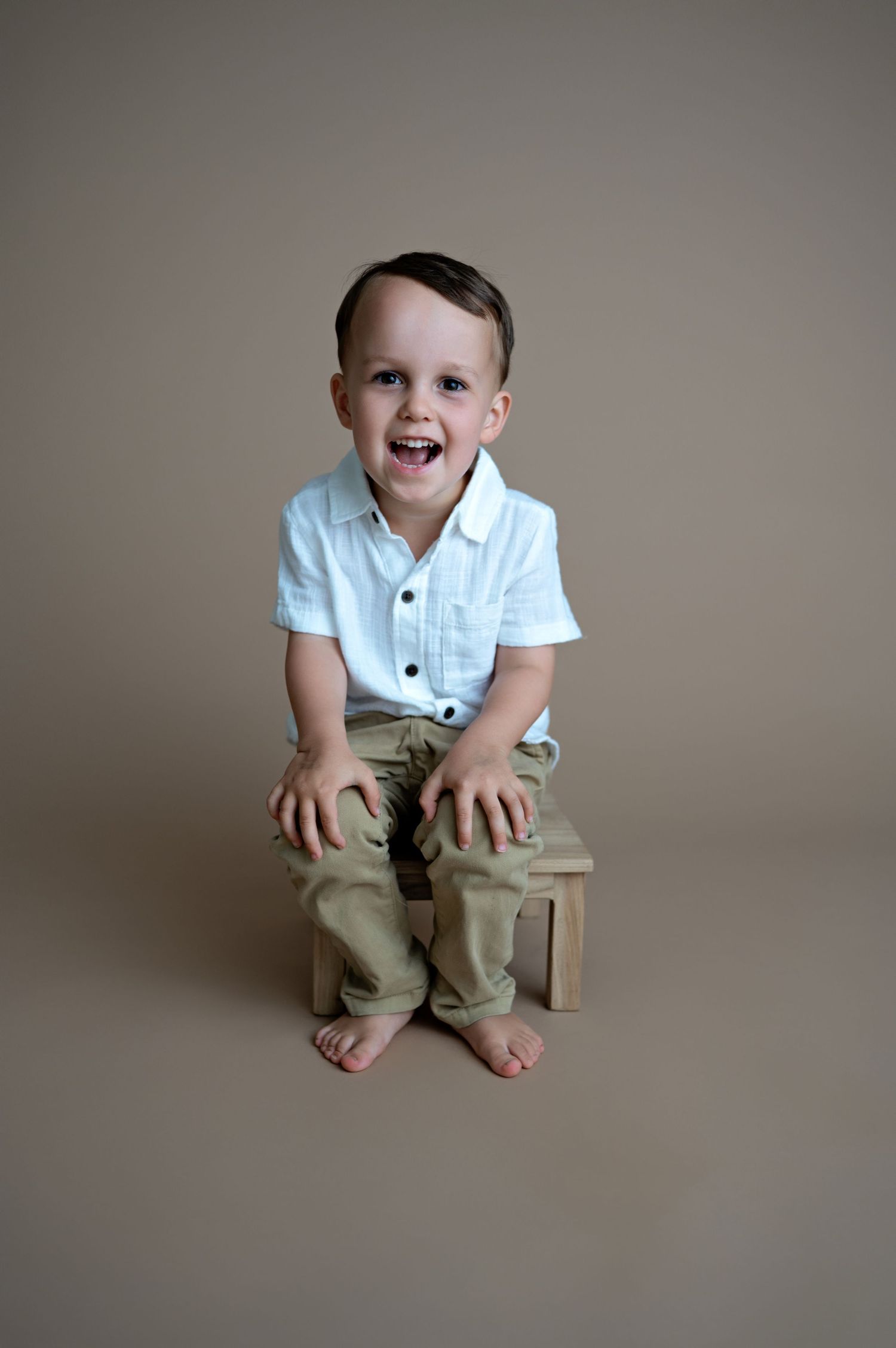 A young child in a white polo shirt and khaki pants sits barefoot on a wooden box against a neutral background.